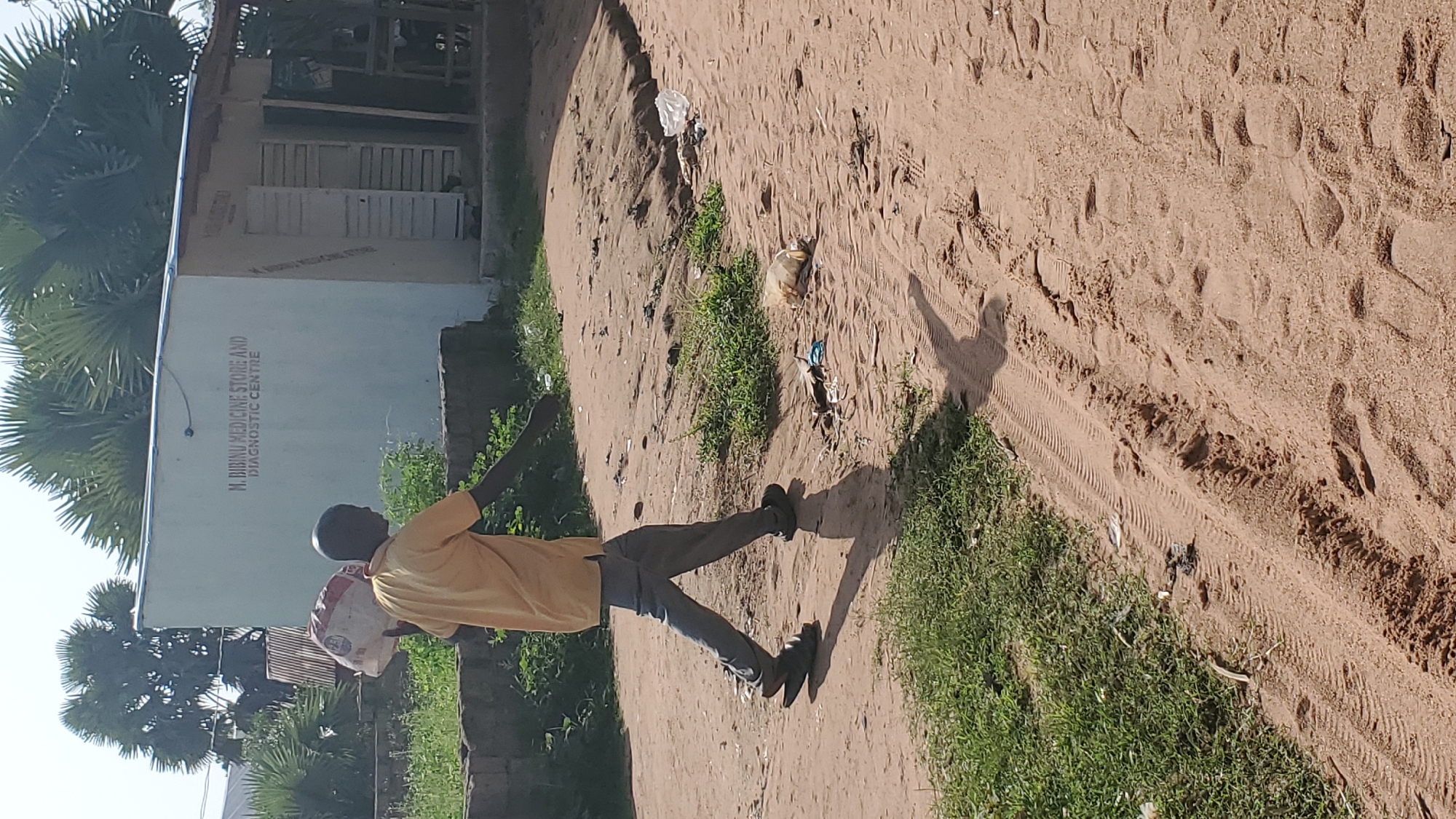 A man is walking across a dirt road in Jalingo, Nigeria, carrying a large, white, plastic bag over his left shoulder. He is wearing a yellow t-shirt and grey trousers, with black sandals on his feet. The man is walking from left to right in the frame. Behind him, a white building with "M. BIBINU MEDICINE STORE AND DIAGNOSTIC CENTRE" written on it in dark letters is visible. The area appears to be dusty and there is some sparse vegetation along the side of the road. Trash is scattered on the ground in front of the building.