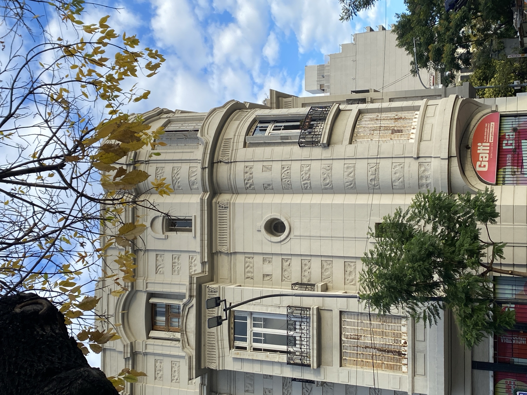The image shows a low-angle view of a multi-story building in Buenos Aires, Argentina. The architectural style features a white or light-colored facade with ornamental detailing, including decorative panels and curved elements. Windows are visible, some with shutters or blinds, and wrought iron details.
A tree with branches and yellow leaves frames the left side of the image, suggesting it's autumn. Another tree with green foliage is on the right.
A red sign with the word "Gatti" is attached to the building's right side, indicating a business name or advertisement. The sign also includes smaller text in Spanish about ventilation and a website address.
In the background, portions of other buildings and a blue sky with clouds are visible. The street lighting fixture is black.
