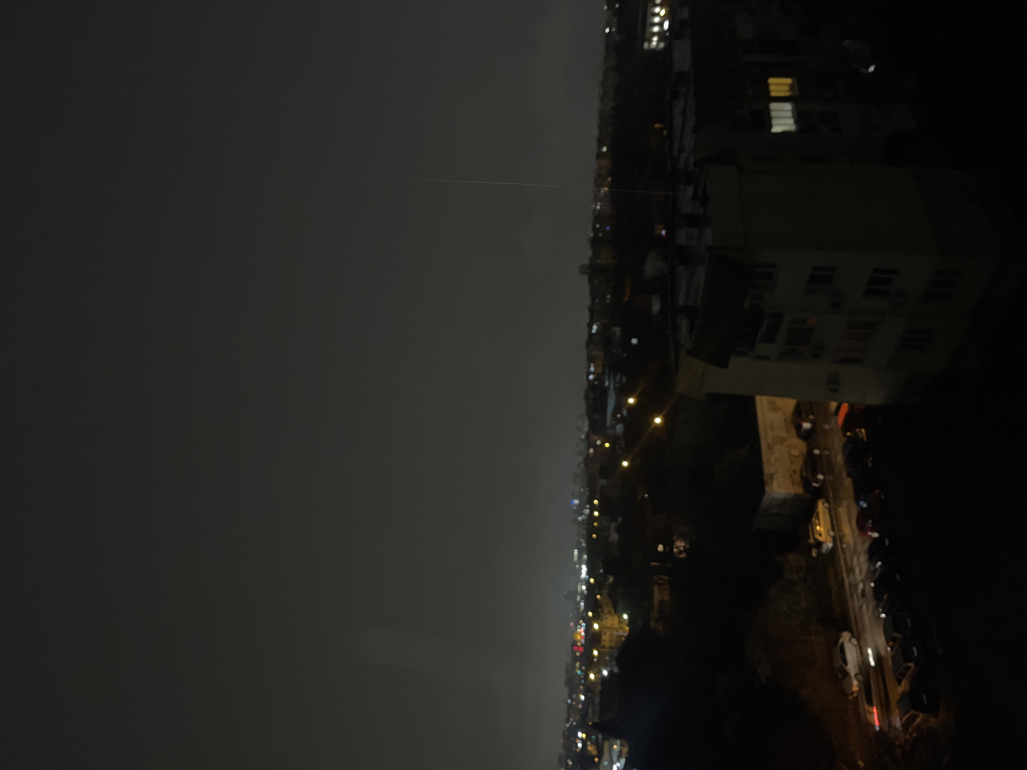 The image depicts an elevated nighttime view of a residential area in Sofia, Bulgaria. The upper half of the frame is dominated by a dark grey, overcast sky, featuring a faint, thin vertical light streak in the upper right quadrant. In the foreground, a wet street is visible, populated by a line of parked vehicles, predominantly dark-colored passenger cars, on the left. Two vehicles, a light-colored sedan with illuminated headlights and a darker vehicle, are in motion on the street. Dark, indistinct vegetation lines the street.
To the right, in the mid-ground, stands a multi-story residential apartment building, light in color, with multiple windows and visible external air conditioning units. Several windows on one facade of this building are brightly illuminated with white and yellow light, indicating active interiors.
The background reveals a sprawling urban landscape characterized by numerous scattered yellow and white lights emanating from buildings and streetlights, extending into a distant atmospheric haze. No individuals are discernible in the scene. The environment suggests an urban residential setting during nighttime, with evidence of recent precipitation on the street surfaces.