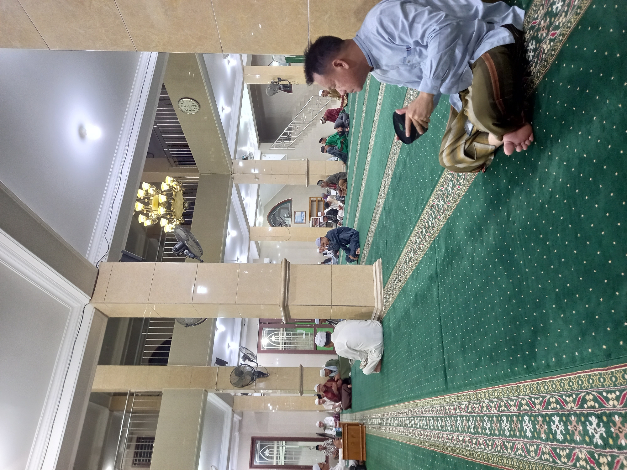 The image depicts the interior of a mosque in Samarinda, Indonesia. The main subject is a man in the foreground, seated on a green carpet with ornate patterns. He is wearing a light blue collared shirt and a striped sarong, and he is holding a black cap in his hands. He is looking to his right, away from the camera.
In the background, a group of men are seated on the same green carpet, facing the same direction as the man in the foreground. They are all wearing traditional Islamic attire, including caps and long robes. Some are engaged in conversation, while others appear to be listening.
The mosque's interior is characterized by large beige pillars, a high ceiling with ornate light fixtures, and a clock on the wall. There are also fans mounted on the walls, suggesting a warm climate. The overall impression is one of a peaceful gathering within a religious space.