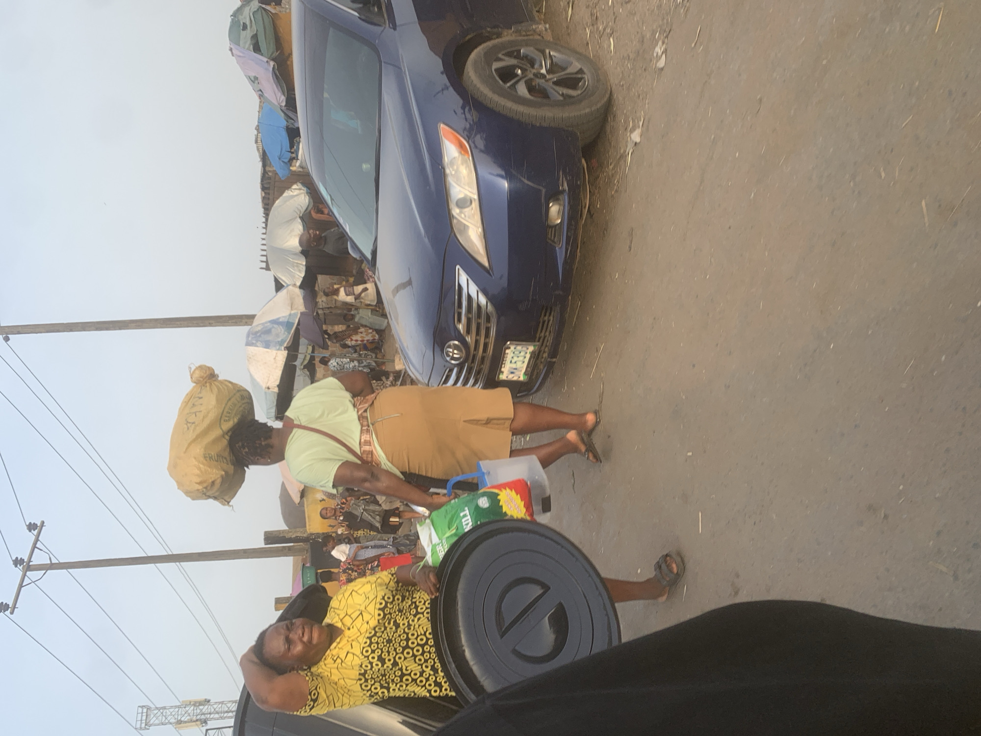 The image captures a bustling street scene, likely a market, under a hazy daylight sky in Fegge, Nigeria. In the foreground, two women are prominently featured.
One woman, positioned towards the lower left, wears a yellow, black, and white patterned dress and holds a large black, circular lid or tray. She appears to be looking upwards and has a slight smile.
To her right and slightly behind her, another woman walks with a large, light brown sack balanced on her head. The sack has visible text including "N.P.F." and "FRUITS." This woman wears a light green short-sleeved top, a tan knee-length skirt, and sandals. In her left hand, she carries a clear plastic container with a blue handle, and a green plastic bag with "TUNS" printed on it.
Behind these individuals, a dark blue sedan, possibly a Toyota Camry, is parked or slowly moving. Part of its front bumper and headlight are visible, along with a license plate that begins with "LAGOS SMK-5". The street surface beneath the car and the people appears to be a mix of dirt and rough asphalt.
In the mid-ground and background, numerous other people are visible amidst market stalls and makeshift shelters, some with umbrellas providing shade. A utility pole with overhead wires is also present on the left side of the frame, extending towards the hazy horizon. The overall impression is one of everyday commerce and activity in a vibrant urban environment.