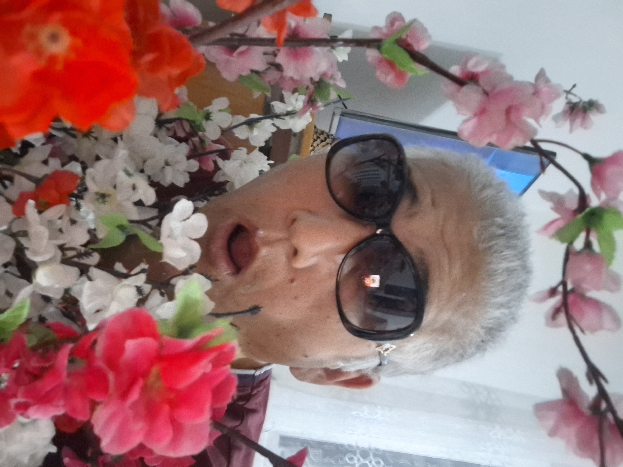 A close-up, selfie-style image shows a person with gray hair wearing large, dark sunglasses and a maroon jacket. The person's mouth is slightly open. The person is positioned in front of a bouquet of artificial flowers, which are pink, white, and red. In the background, a television screen is visible. The scene appears to be indoors, possibly in a home. There is no indication of the city of Almaty, Kazakhstan.