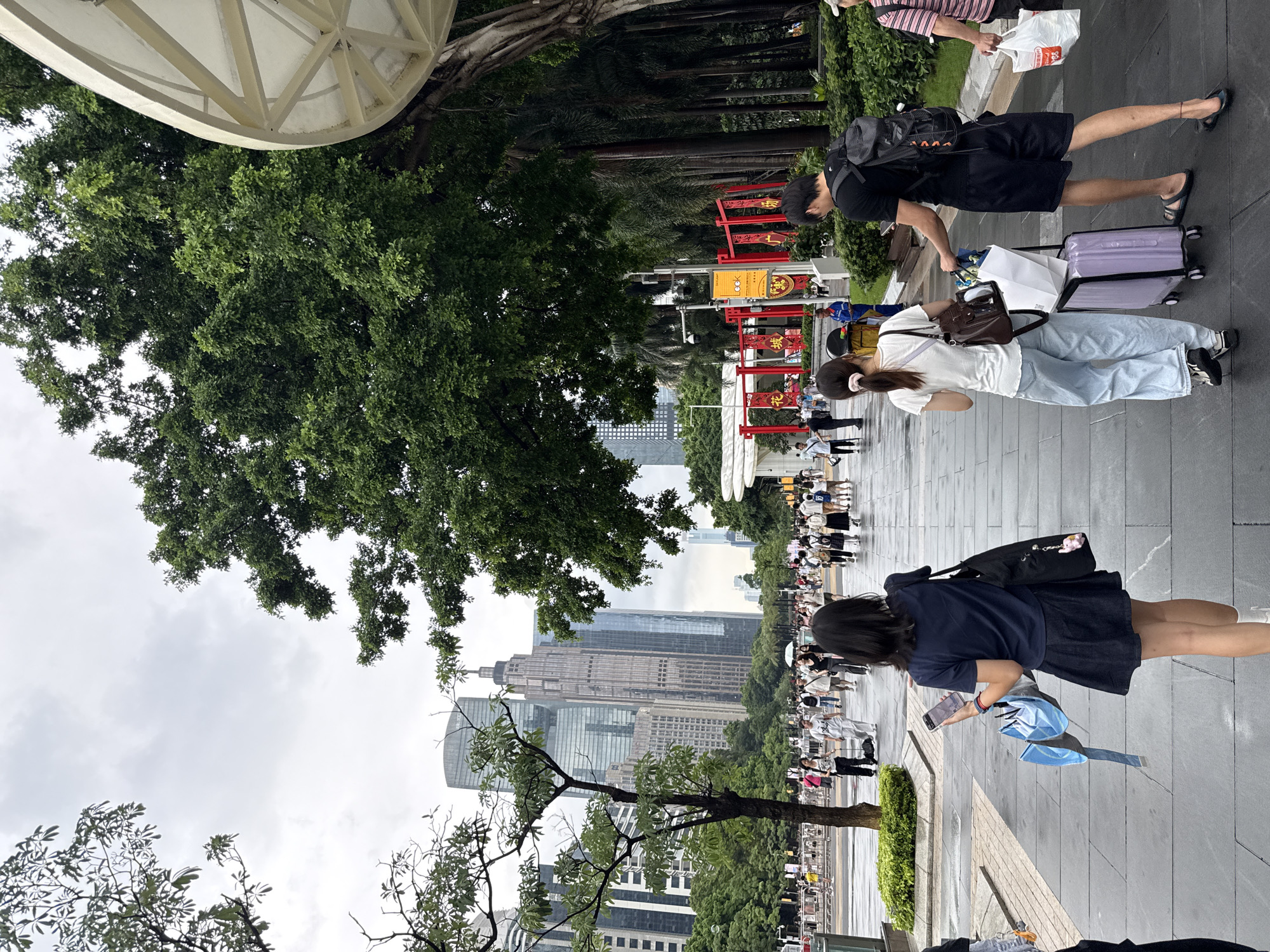 The image captures a street scene in Tianhe District, China. The perspective is from a low angle, looking upwards, with the majority of the image oriented vertically. The foreground is dominated by lush green trees and a portion of a white, modern structure with a ribbed surface.
In the middle ground, people are seen walking along a paved walkway. Some individuals are pulling luggage, suggesting travel. One woman in a white t-shirt and light blue jeans is walking with a brown bag over her shoulder, next to a man in a black t-shirt and black shorts, who is also carrying a backpack and pulling a purple suitcase. Further down the walkway, more people are visible, some in groups. Red and yellow decorative arches, possibly for a festival or event, are spaced along the pathway.
The background features a dense line of palm trees, beyond which rise numerous tall, modern buildings, characteristic of a city skyline. The sky is overcast and appears to be a dull, cloudy day. The overall atmosphere suggests a busy urban environment with people going about their daily activities or traveling. No specific time of day is clearly indicated, but the overcast weather suggests it could be daytime. No visible text is discernable.