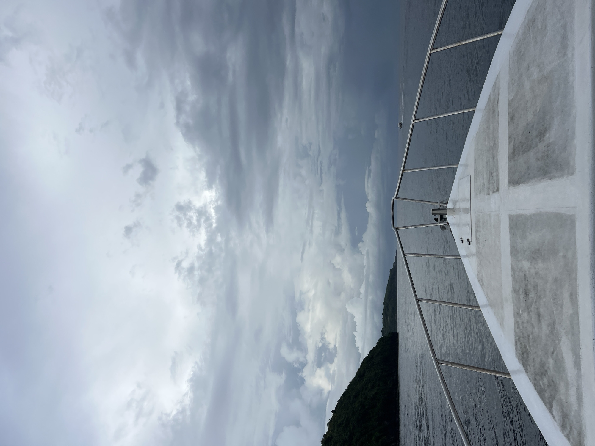 The image captures a maritime scene from the perspective of a boat on open water, likely off the coast of Nusa Penida, Indonesia. The white deck of a boat or yacht, featuring grey textured non-slip patches and gleaming metal railings, is prominently visible on the right side of the frame. The calm, dark grey-blue water stretches across the majority of the lower and central parts of the image, reflecting the cloudy sky above.
In the distance, a lush green island covered with dense tropical vegetation rises from the water on the left, grounding the scene geographically. Further out on the water, a small, dark speed boat can be seen, leaving a faint white wake behind it as it travels across the horizon.
The sky is heavily overcast with a thick blanket of grey and white clouds, suggesting cloudy weather, possibly before a rain shower or during an otherwise subdued day. Despite the heavy clouds, the general illumination indicates it is daytime. The overall mood is one of tranquil solitude on the water. There are no people visible, nor is there any discernible text within the image.