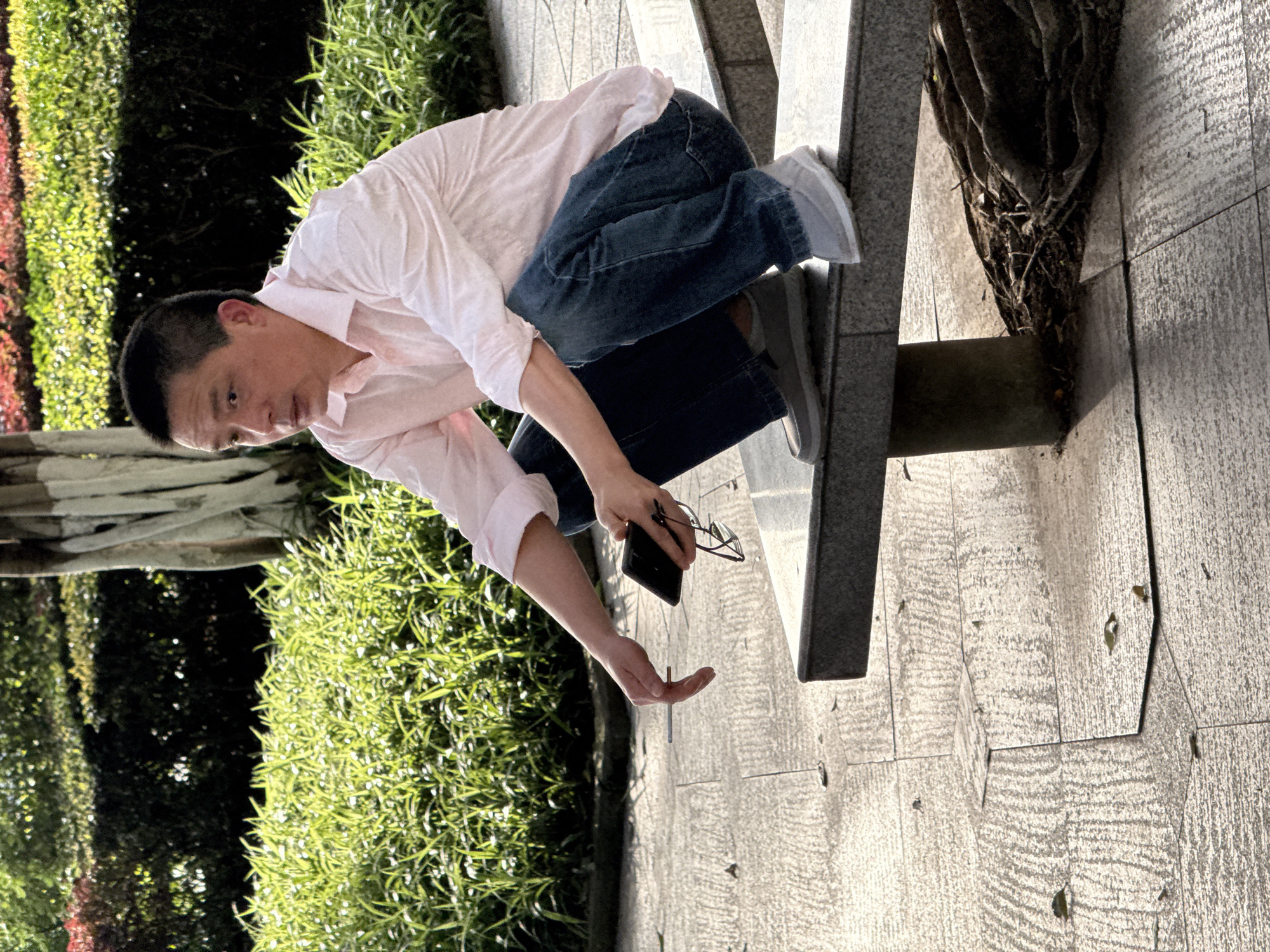 The image depicts a man in an outdoor setting, likely a park or public pathway in Jiangjin, China. He is wearing a light pink long-sleeved shirt, dark blue jeans, and grey sneakers with white soles. The man is captured in a bent-over posture, with his right knee resting on a light grey concrete structure, possibly a planter or bench. His head is tilted downwards, and his gaze is directed towards the ground, indicating intense focus on something below him.
His left arm is extended forward with an open palm, suggesting he is reaching for or pointing at an object on the ground. In his right hand, he holds a black smartphone and a pair of dark-framed eyeglasses.
The background features dense green foliage and bushes, with some reddish plants visible further behind. The ground is paved with textured grey and darker grey tiles, arranged in a pattern of squares and hexagons. To the right of the concrete structure, a gnarled tree trunk or root system is visible. The scene is brightly illuminated by natural sunlight, indicating a clear, sunny day. The man's expression appears focused and concentrated. There is no visible text in the image.