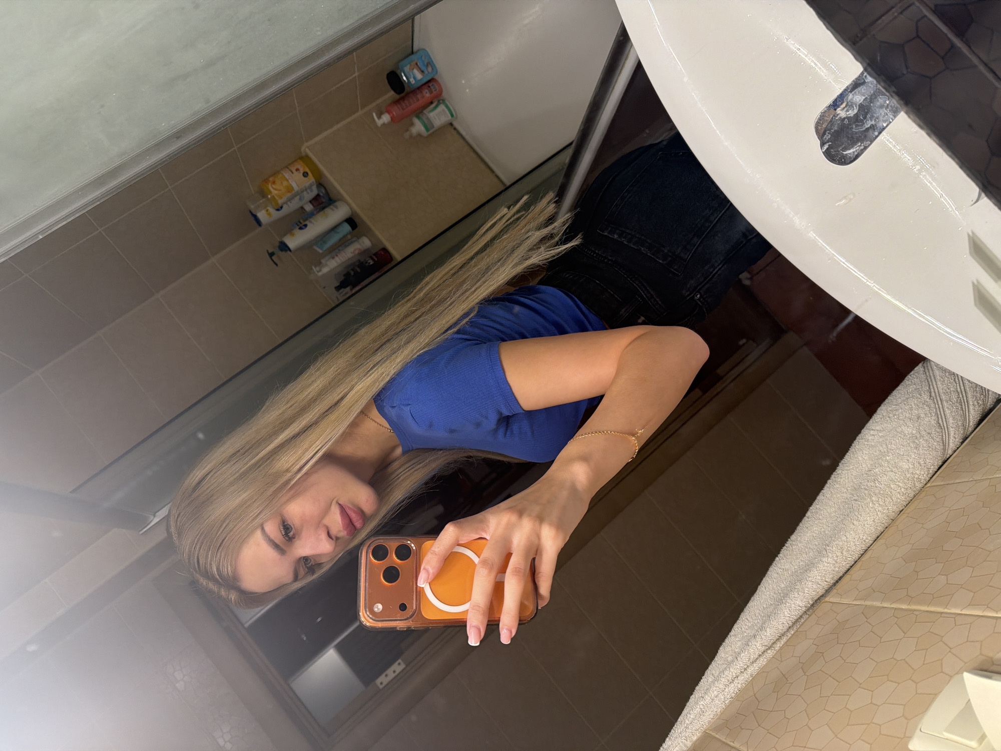 A young woman with long blonde hair takes a mirror selfie in a bathroom. She is wearing a blue ribbed t-shirt and dark wash jeans. She is holding a bright orange-cased smartphone, and a delicate gold bracelet is visible on her wrist. The bathroom features beige tiled walls, a white sink, and a bathtub in the background, where several bottles of toiletries are arranged on a shelf. The overall impression is of a casual, private moment captured in a domestic setting.