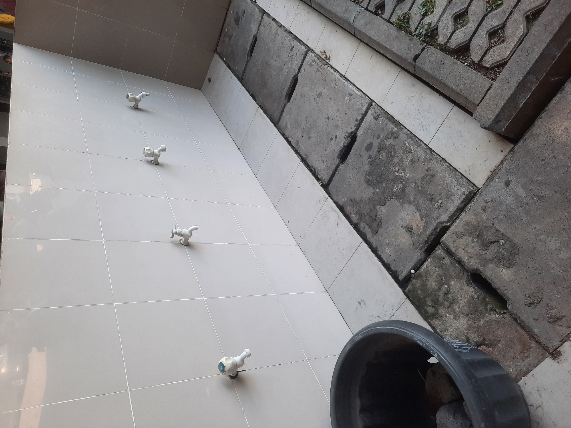 The image depicts an outdoor communal washing area, likely in North Jakarta, Indonesia. The scene features a tiled wall with four white faucets. In front of the wall, a row of concrete slabs covers a drain. To the left, a large black plastic tub sits on a tiled floor. The ground is a mix of tiles and concrete, with some weeds growing in the cracks. The overall impression is one of utilitarian outdoor infrastructure, possibly found in a public park or a community facility. There are no people visible in the image.