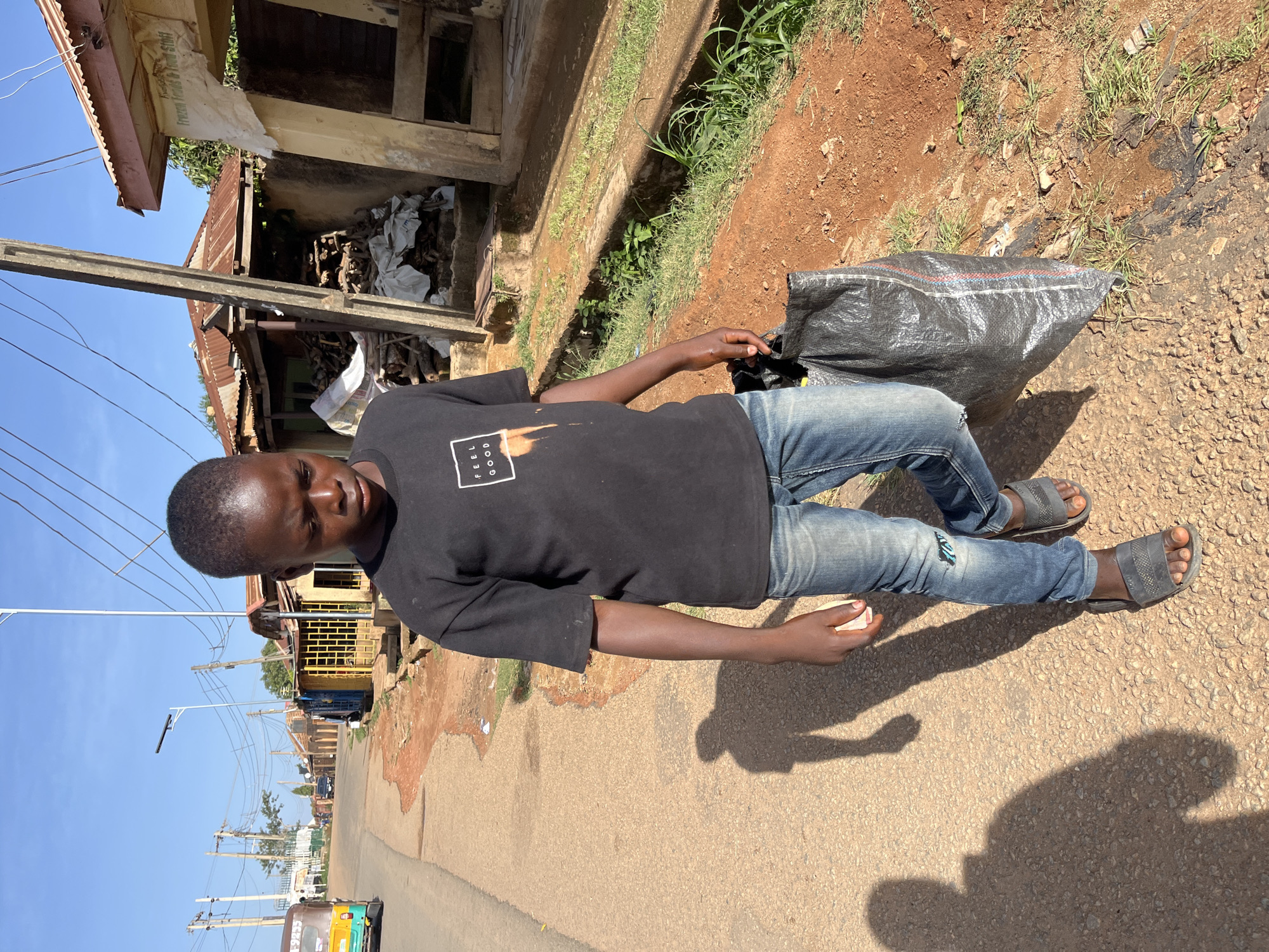 The image features a young male standing on the side of a paved road under a clear, bright blue sky. He has dark skin and short dark hair, looking directly at the camera with a neutral expression. He is dressed in a black short-sleeved t-shirt with a white square logo displaying "FEEL GOOD" and ripped blue jeans, paired with dark grey slide sandals. In his left hand, he holds a large black woven plastic sack with visible red and blue stripes; in his right hand, he holds a small, light-colored rectangular object.
Strong shadows cast on the ground suggest it is sunny, likely mid-morning or early afternoon. The road surface is a mix of asphalt and gravel, transitioning to a reddish-brown dirt shoulder with sparse green grass and a concrete drainage ditch. Beyond the ditch, several aged buildings with corrugated metal roofs and wooden supports line the street. One building has a faded banner partially visible, which reads "KUMSAL" and "Frozen Foods & Your Stuff!".
The background reveals a street scene with numerous utility poles, overhead power lines, and newer streetlights. In the distance, a brightly colored auto-rickshaw (Keke Napep) with the numbers "9235" partially visible, is parked on the road. The setting depicts a bustling urban or semi-urban environment.
