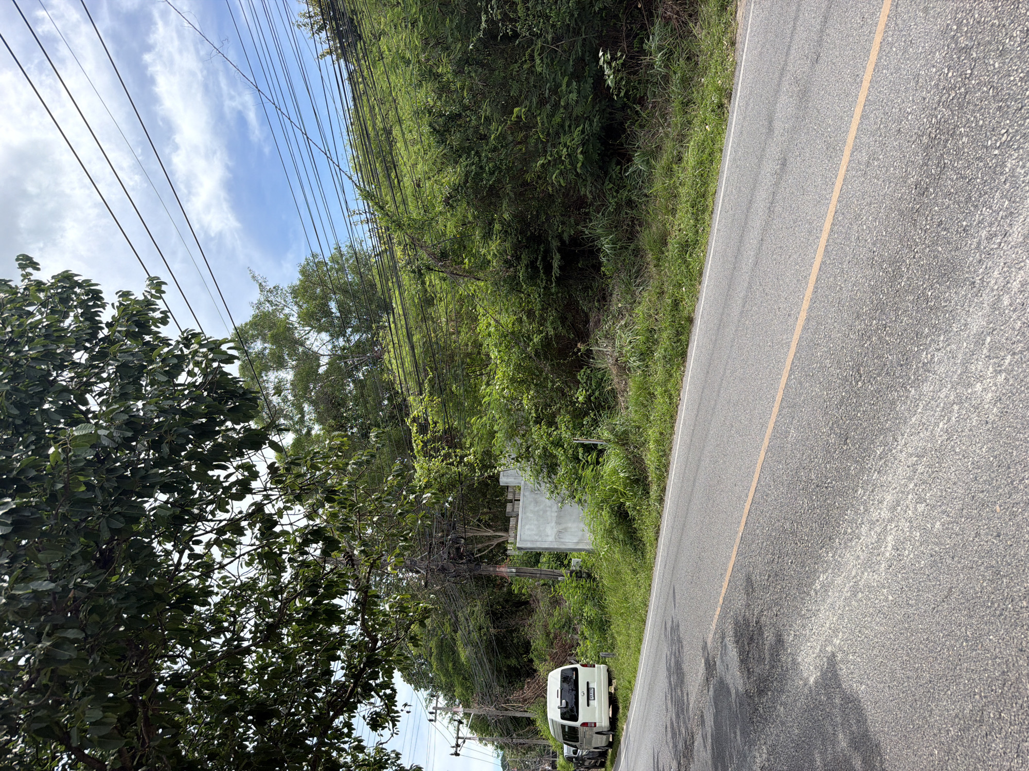 A white van with a license plate ending in "8129" is parked on the side of a paved road in Choeng Mon, Thailand. The van is positioned to the left of the frame. The road has a single yellow line down the center and appears to have some patches and wear. To the right of the road, there is a steep embankment covered in lush green vegetation and trees. Electrical wires are strung overhead and across the hillside, supported by utility poles. A section of a light-colored concrete structure is visible on the embankment, partially obscured by foliage. The sky above is partly cloudy.