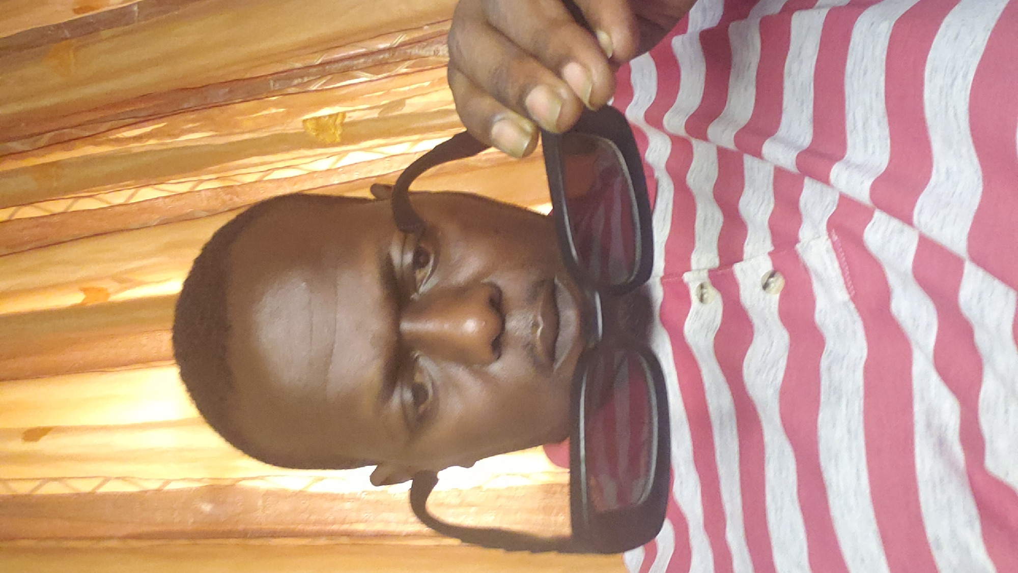 An individual, possibly a man, is holding a pair of black-framed sunglasses in front of his face. He is wearing a red and white striped shirt. The background appears to be a room with what seems like curtains or fabric draped on the wall. There is no clear indicator for the city of Narayi, Nigeria.