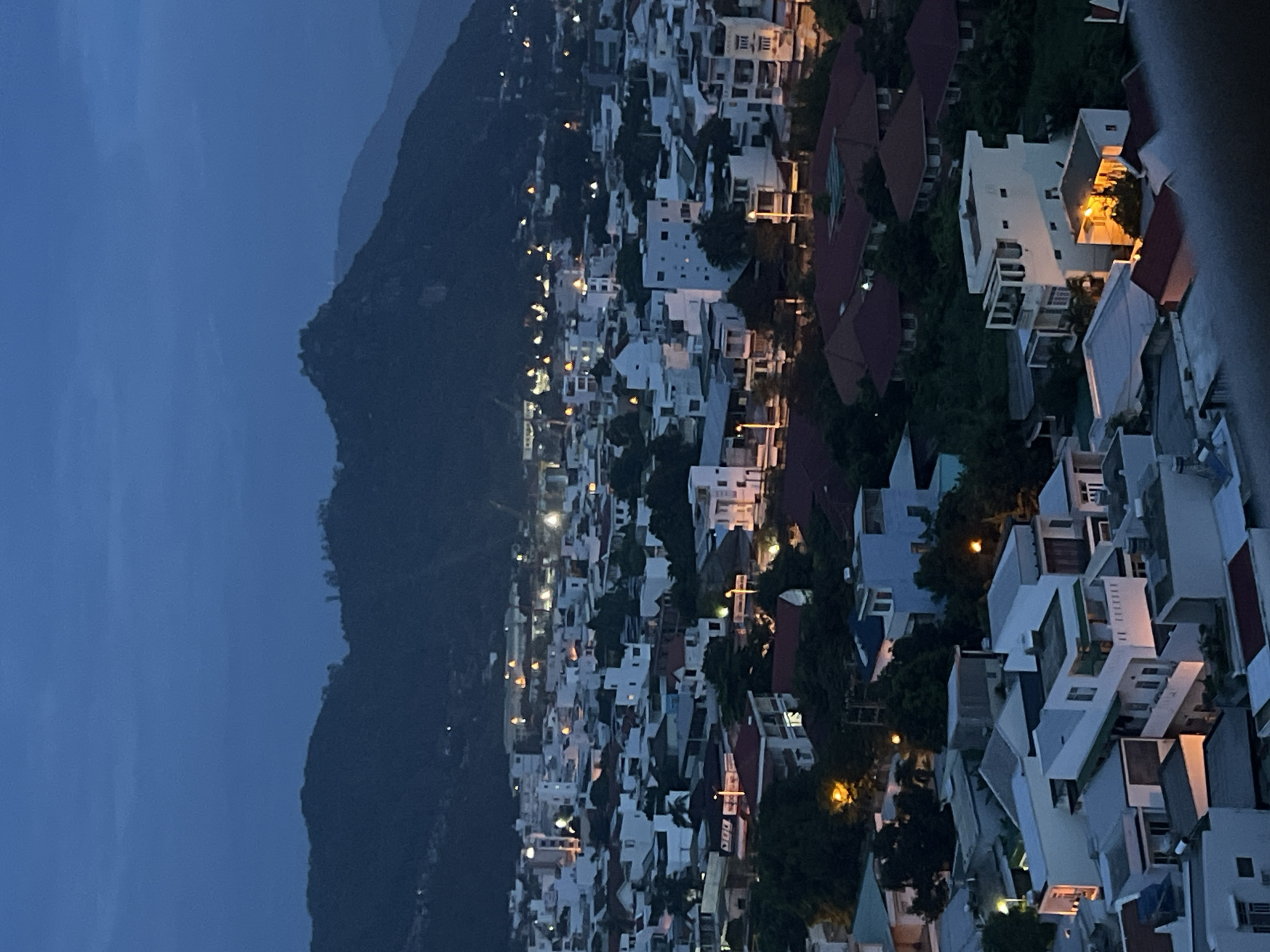 The image captures a panoramic view of a densely populated residential area at dusk or early night in Bac Nha Trang Ward, Vietnam, set against a dramatic mountain backdrop.
On the left side of the frame, a dark mountain range looms, its contours visible against the deep blue twilight sky. The rest of the image is dominated by a sprawling urban landscape, featuring numerous white and light-colored buildings. These structures display a mix of architectural styles, with many having flat roofs and others sporting distinct red-tiled roofs. The buildings are multi-storied and appear closely packed, suggesting a hillside development or a dense urban environment.
Artificial lights, predominantly warm yellow and orange, glow from within the houses, balconies, and street lamps, casting pockets of illumination across the scene. These lights create a stark, inviting contrast with the cool blue tones of the sky and the dark mountain. Dark green trees and foliage are interspersed among the buildings, indicating lush vegetation throughout the residential area. The photograph is taken from an elevated vantage point, possibly from a higher building, looking down over the city. A blurred, grey element on the extreme right edge suggests a railing or part of the immediate foreground structure.
There are no visible people or specific activities taking place; the image is a static cityscape. No text is discernible. The overall impression is one of a peaceful urban night scene, highlighting the architectural style and the natural beauty of the surrounding landscape as the city transitions from day to night.