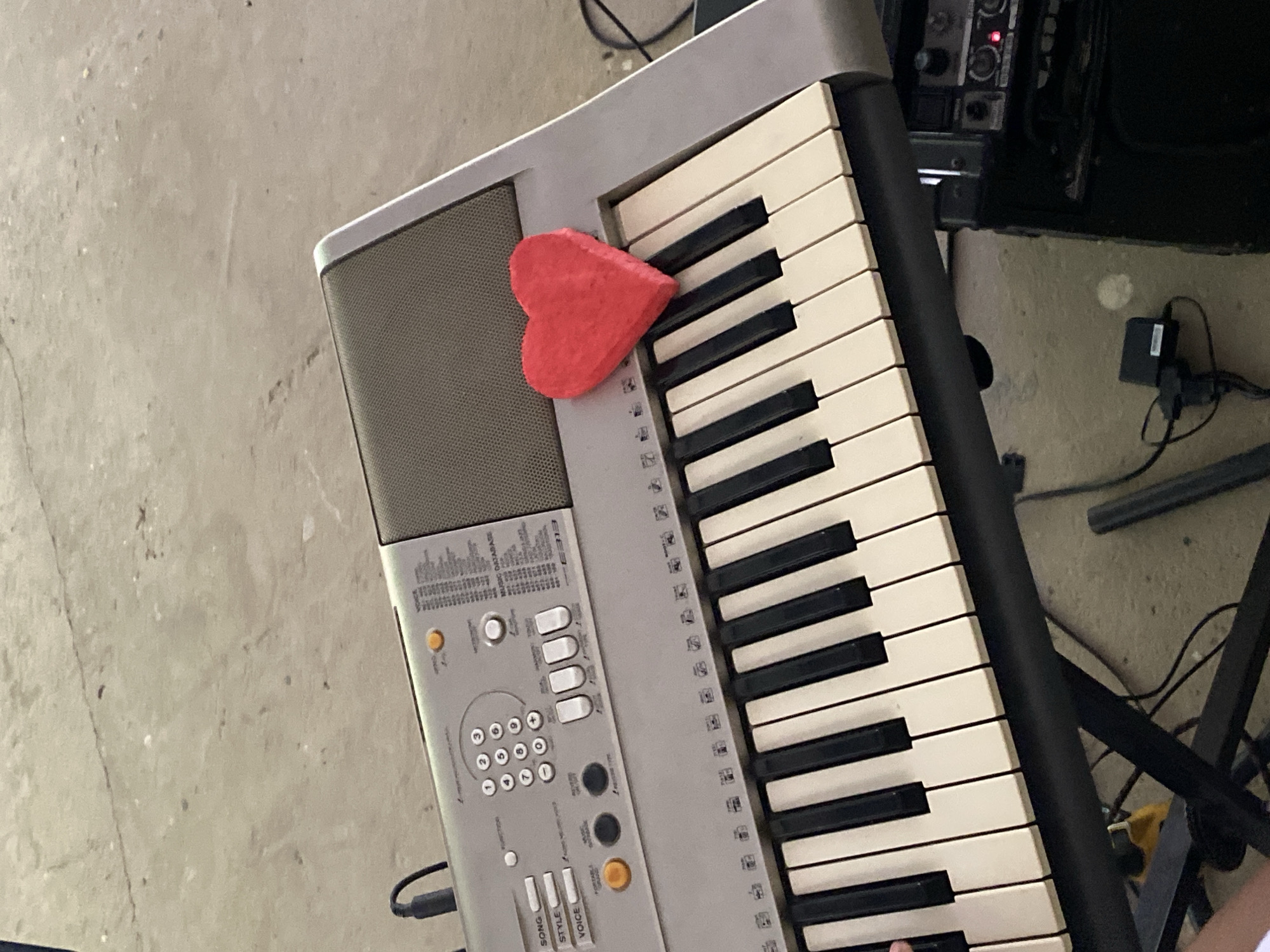 A digital keyboard is positioned at an oblique angle. A red, heart-shaped object rests on the left side of the keyboard, partially covering the speaker grill and some of the keys. The keyboard is gray with black keys, and the keys are ivory. The background consists of a gray concrete floor and some audio equipment, including a black amplifier. A tangled arrangement of cords, wires, and a black power adapter are on the floor. Part of a black metal keyboard stand is also visible on the right side of the image. The city of Tinaquillo and Venezuela are not discernable.