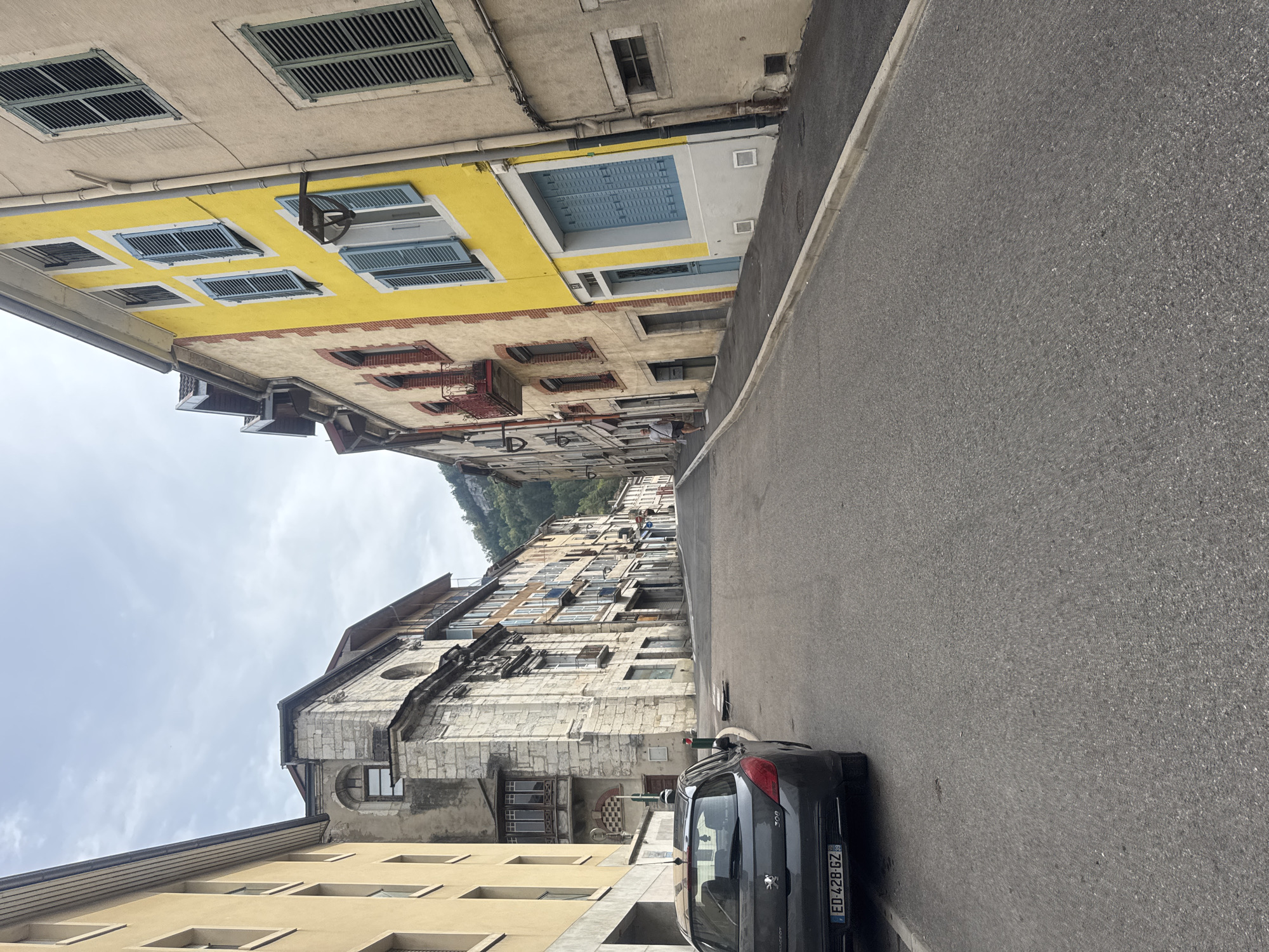 A street scene in Saint-Claude, France, depicts a narrow, uphill road lined with multi-story buildings. On the left, a dark gray Peugeot 308 hatchback with license plate ED-428-GZ is parked. The buildings on both sides of the street exhibit a mix of architectural styles, with varying colors and window designs. The building on the right is particularly striking with a bright yellow facade on its upper floors, featuring blue shutters on several windows. The street is paved with asphalt, and a sidewalk runs along the right side of the road. The sky is overcast with clouds. Further up the street, a person is walking away from the camera. In the background, a lush green hillside is visible, suggesting the town is situated in a valley or mountainous region.