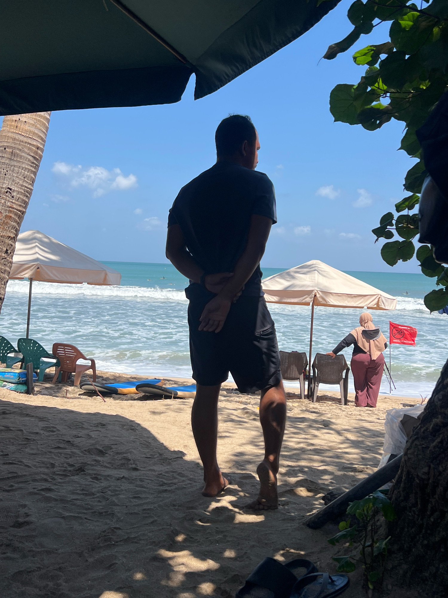 The image depicts a sunny day at Kuta Beach, Indonesia, characterized by a sandy shore, ocean waves, and clear blue skies dotted with clouds. In the foreground, a man stands with his back to the camera, wearing a dark t-shirt and shorts. He has his hands clasped behind his back and is looking out towards the ocean. To his left, a palm tree trunk is visible.
In the mid-ground, several beach chairs are arranged on the sand. Two surfboards are also leaning against each other. A large beach umbrella shades a portion of the scene. Further in the background, a woman wearing a headscarf and a long, flowing outfit stands near another beach umbrella, holding onto a chair. A red flag with a white symbol is planted in the sand to her right, indicating a warning or prohibition.
The ocean is visible with moderate waves rolling towards the shore, and several surfers can be seen in the distance. The overall atmosphere suggests a relaxed beach setting, likely a tourist area in Kuta, known for its surfing and vibrant atmosphere. The image is taken from under a large, dark green umbrella, with the edge of its fabric visible at the top of the frame. Foliage from a tree can be seen on the right side of the image, adding to the natural environment of the beach.