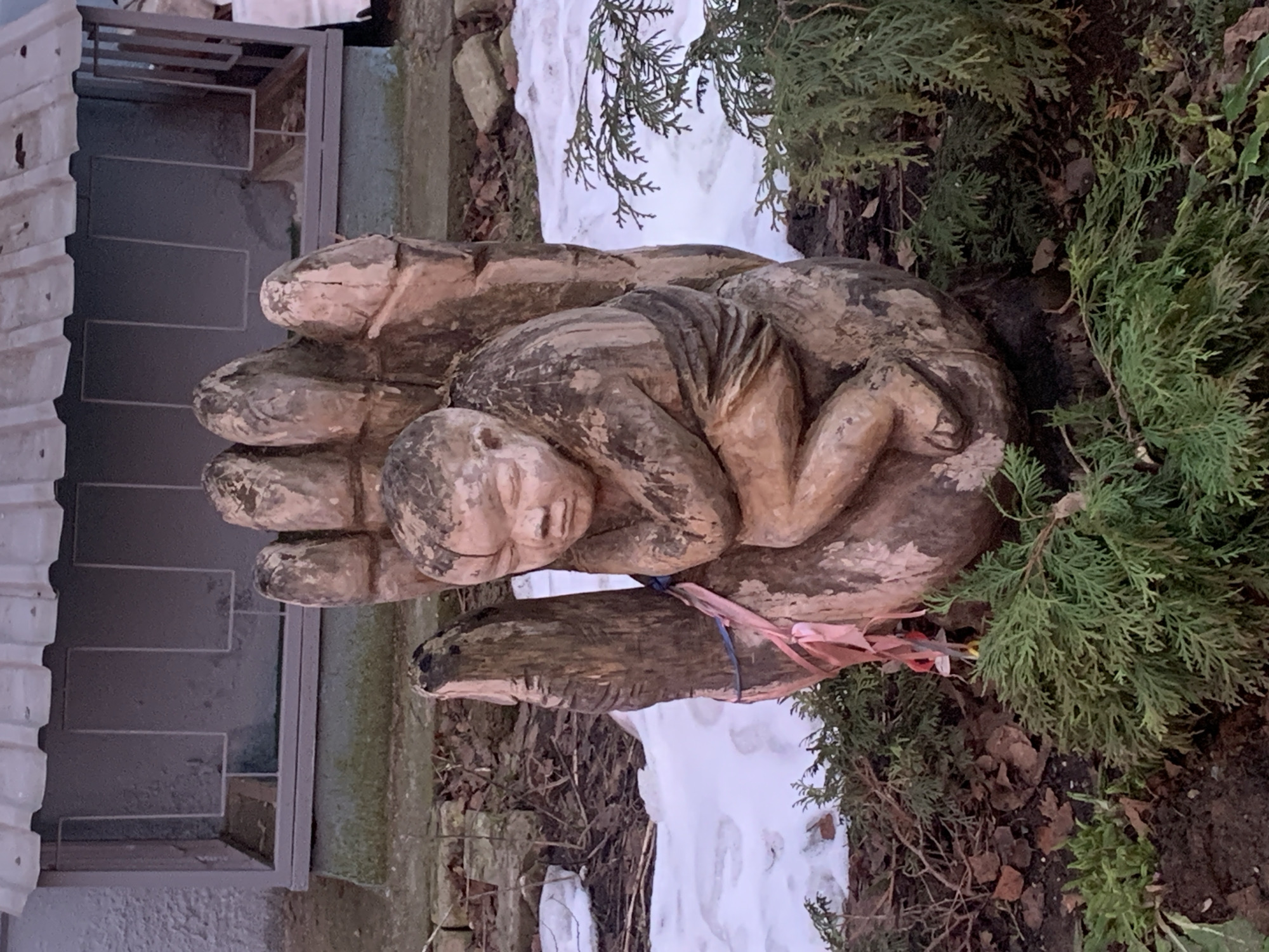 A wooden sculpture of a hand holding a human figure is located near a building with steps. The sculpture is surrounded by evergreens and patches of snow. The location is likely Chaikovsky, Russia. The figure is lying down in the hand, suggesting a gesture of protection or support. A light blue and pink ribbon is tied around the hand sculpture.