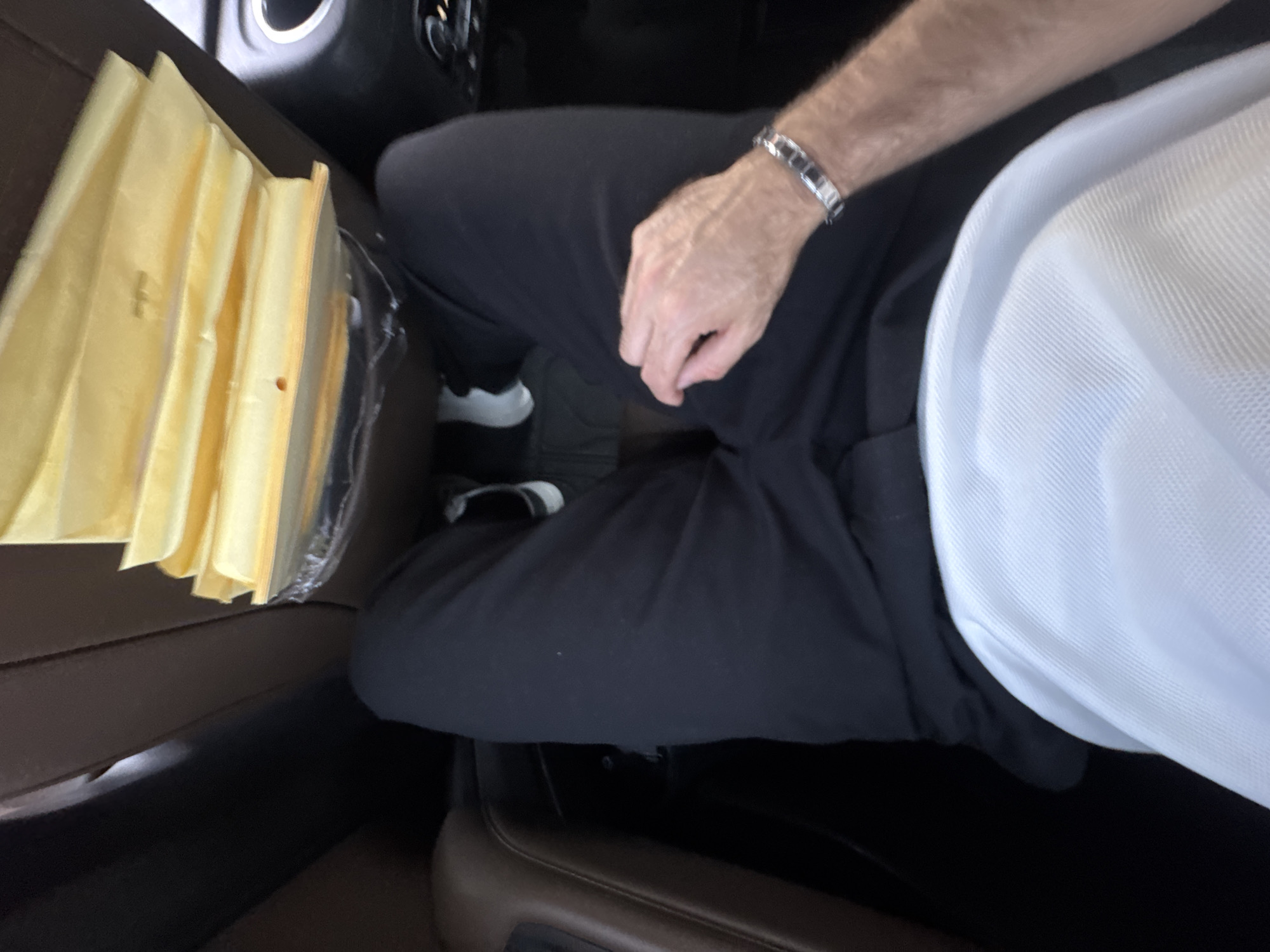 The image captures a top-down view of the interior of a vehicle, likely a car, featuring a person and a stack of objects. A person is seated, with their lower torso and legs, clad in dark pants, visible. Their right arm and hand are visible, resting on their lap, and they are wearing a metallic, possibly silver-toned, link bracelet on their wrist. A portion of a white, textured fabric garment, likely a shirt or undershirt, is visible on their upper right.
To the left of the person, a stack of numerous thin, rectangular, yellow-beige items is present. These items appear to be made of a slightly translucent material, possibly paper envelopes, bags, or thin foam sheets, and are bundled together, with some clear plastic visible around them. The items show some creasing and a few small perforations. The immediate setting around the person and objects consists of dark vehicle interior elements, including what appears to be part of a car seat or console upholstered in a brown material, and darker flooring or panels. The lighting suggests an indoor setting, consistent with a vehicle interior, but the time of day is not discernible. No specific location cues are visible within the image itself, though the context provided indicates Mexico City, Mexico. There is no visible text. The activity depicted is simply sitting within the vehicle.