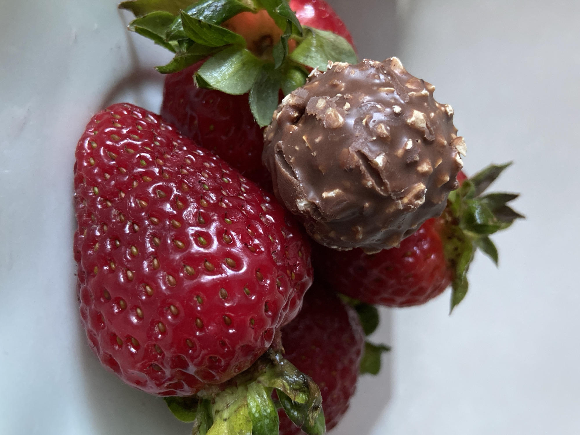 A close-up, top-down shot captures a tempting arrangement of fresh strawberries and a chocolate truffle against a soft, light background. The primary subjects are several ripe, red strawberries, characterized by their glossy skin and visible yellow-green achenes (seeds) embedded across their surface. Most of the strawberries retain their vibrant green calyxes (stems and leaves), suggesting freshness.
Nestled among the strawberries is a single, round chocolate truffle, resembling a Ferrero Rocher. It is coated in a textured layer of milk chocolate mixed with crushed nuts, likely hazelnuts. The truffle appears rich and inviting, contrasting with the bright, natural redness of the fruit.
The items are arranged in a cluster, partially overlapping, and fill the majority of the frame. The background is an out-of-focus, plain white or very light gray, which effectively highlights the vibrant colors and textures of the subjects. Reflections are visible on the glossy surfaces of the strawberries, indicating direct lighting.
No specific activity is taking place; the items are presented statically, possibly awaiting consumption or as a still-life composition. The overall impression is one of fresh, sweet indulgence. There is no visible text in the image. The setting appears to be indoors, likely a simple domestic environment, given the neutral background.