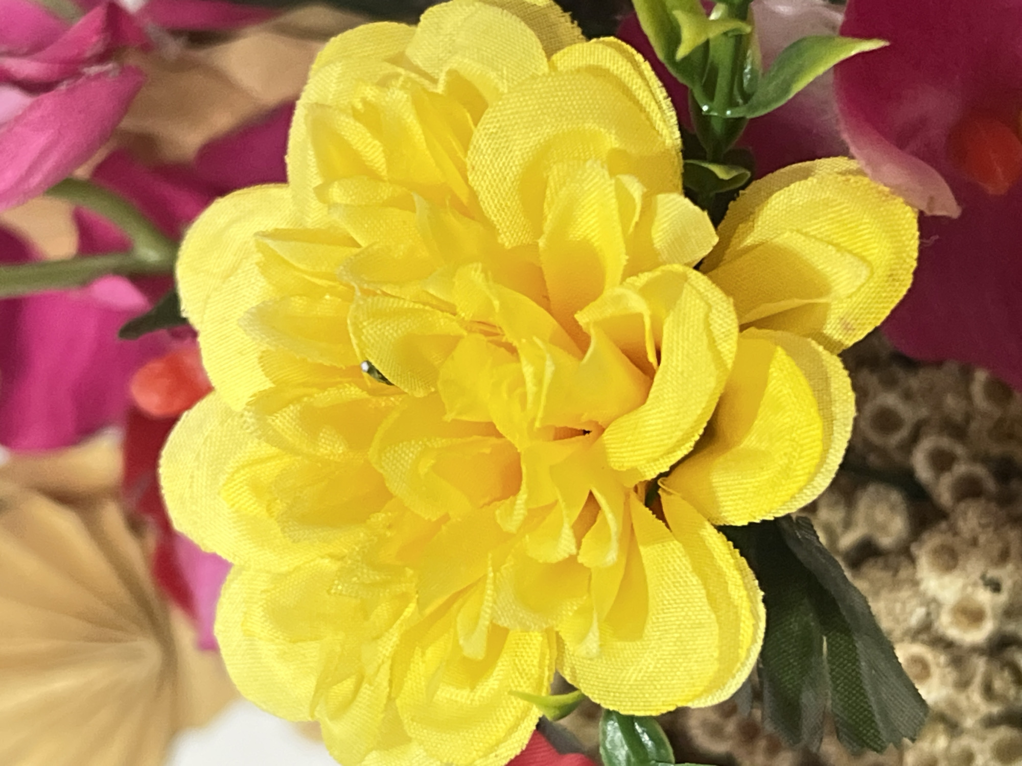 A close-up, eye-level, vertical shot showcases a vibrant artificial yellow flower as the central subject. The flower has numerous layered petals, creating a full and textured appearance. It is part of a larger floral arrangement, with hints of pink and tan flowers visible in the blurred background. Small green leaves are interspersed among the petals. The overall lighting suggests an indoor setting, possibly a home or a shop in Denpasar, Indonesia, given the context provided. There are no people or actions depicted in the image; it is a static arrangement of decorative flowers.