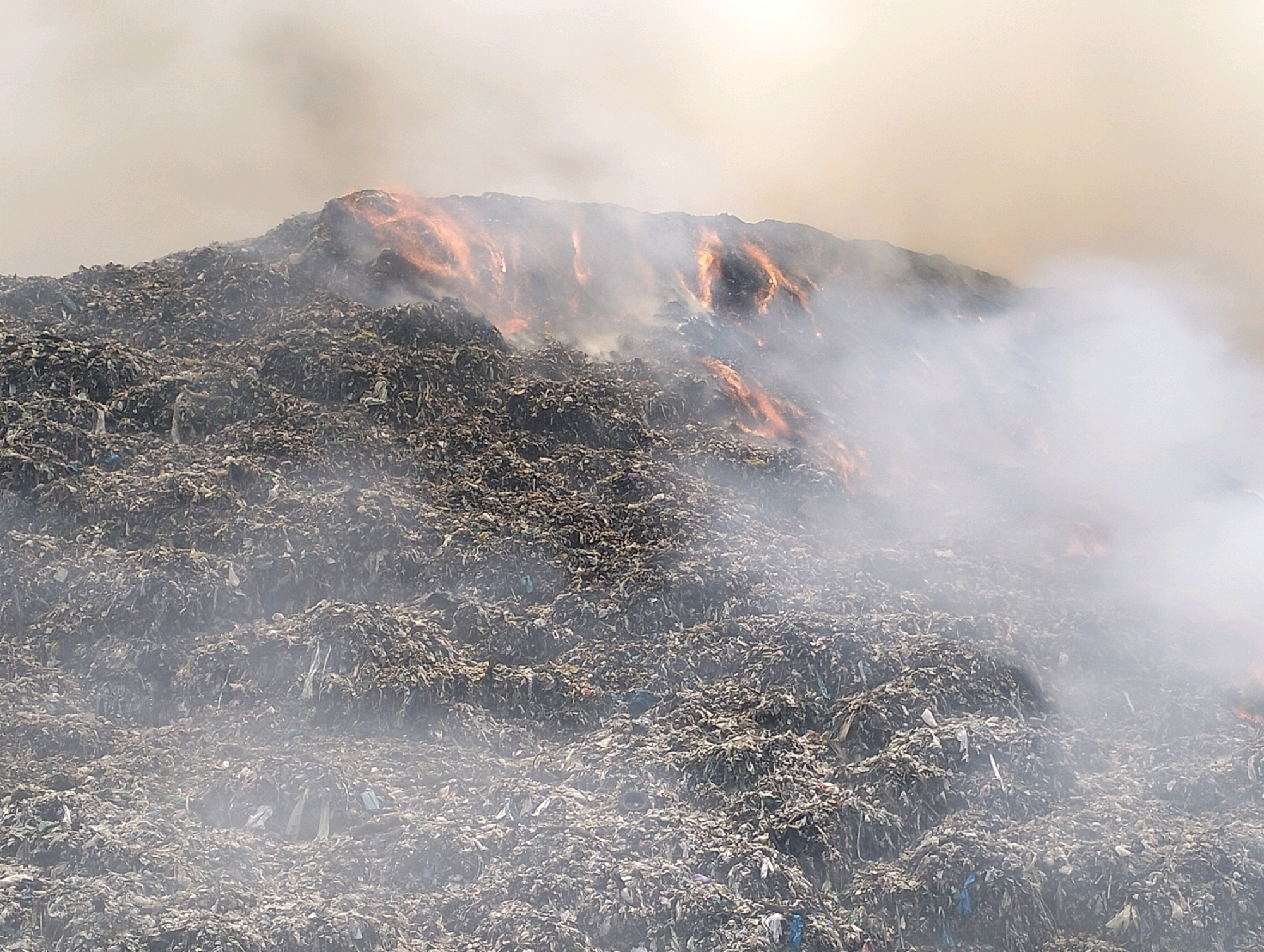 The image depicts a large, smoldering landfill in the city of Surakarta, Indonesia. The landfill is a massive pile of debris, appearing to be primarily household waste, including plastic bags, textiles, and other refuse. Several areas within the landfill are actively burning, indicated by visible flames and glowing embers that cast an orange hue on the surrounding material. Thick plumes of white and grey smoke rise from the burning sections and billow across the landscape, obscuring parts of the landfill and the sky. The overall scene is one of environmental degradation and potential hazard due to the uncontrolled burning of waste. There are no visible people or specific structures identifiable within the frame, only the vast expanse of the garbage dump.