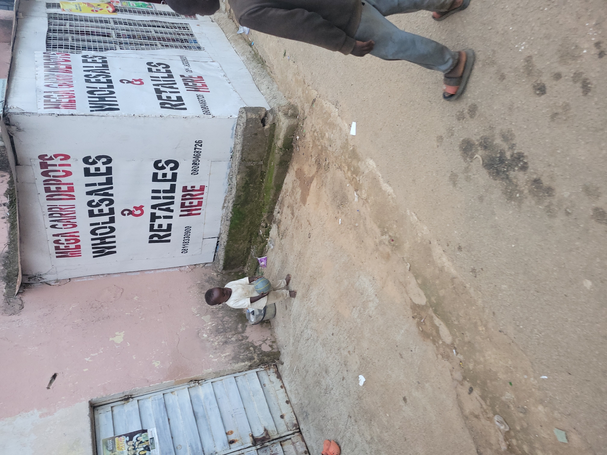 This is a medium shot, eye-level, outdoor scene in Jos, Nigeria. A young boy, appearing to be around 3-4 years old, stands barefoot on a dirt and concrete ground in front of a commercial building. He is wearing a light-colored t-shirt and trousers and is holding a blue and green ball. To the right of the frame, another child, an older boy, walks away from the camera, wearing a brown sweater and blue trousers, with sandals on his feet. The building behind the younger boy is painted a pale pink on the left and white on the right, with a large sign advertising "MEGA GARRI DEPOTS WHOLESALES & RETAILS HERE" in bold red and black lettering. The building also has a metal grate on the right side, suggesting it is a shop. The ground is a mix of packed dirt, concrete, and some scattered debris. The overall environment appears to be an urban street or market area in Jos, Nigeria, characterized by its functional, no-frills commercial spaces and the presence of children.