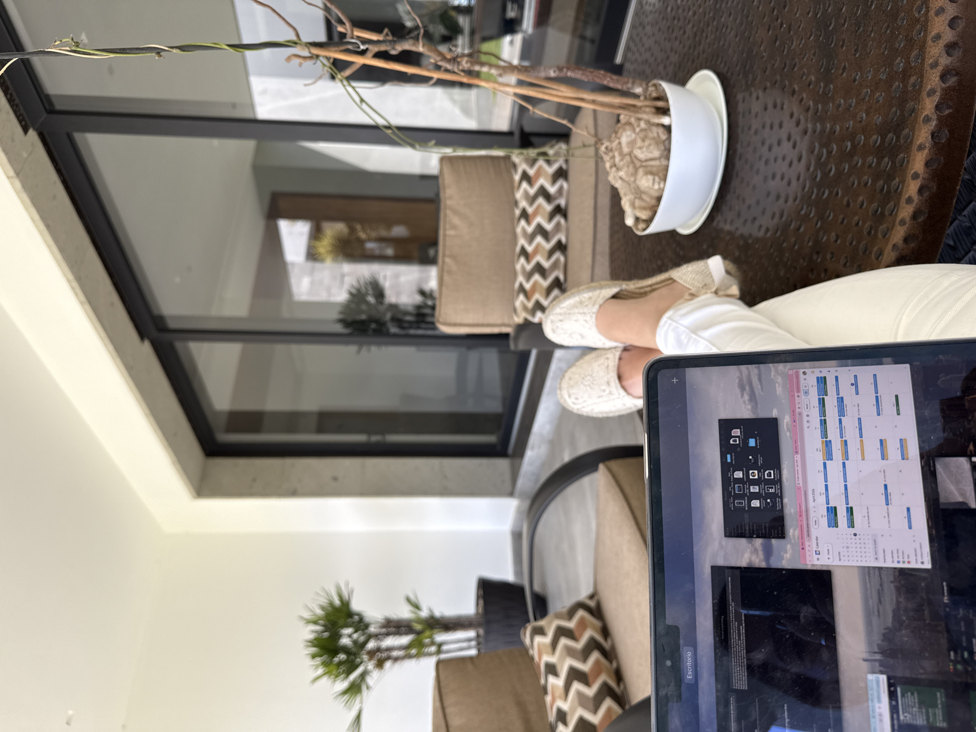 The scene depicts a person working on a laptop, with feet propped up, potentially on a balcony or patio. The laptop screen displays what appears to be a file manager and a calendar application. The environment includes outdoor seating with cushions, a decorative plant in a white pot, and a view through a large window or door revealing outdoor greenery. Based on the location information provided (San Bernardino Tlaxcalancingo, Mexico), the architectural style and plant types (which seem to include palms), it is likely a residence with a modern design aesthetic.
