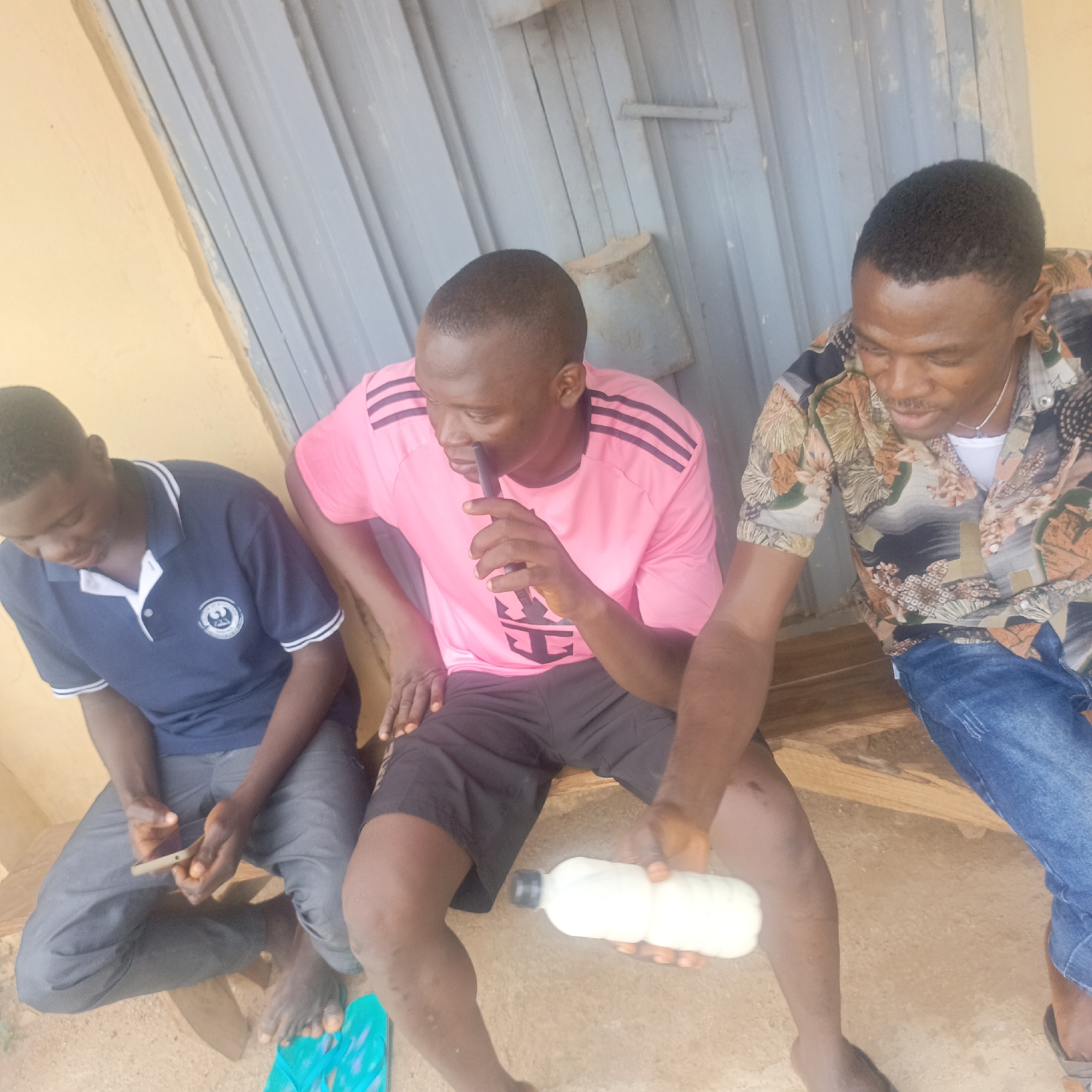 Three young men are sitting on a wooden bench outdoors in Akwanga, Nigeria. The man on the left wears a navy blue polo shirt and grey pants, and holds a mobile phone in his hands. The man in the middle wears a pink t-shirt with black stripes and black shorts, and is holding a black object to his mouth, possibly an e-cigarette or vape. The man on the right wears a patterned shirt and blue jeans, and is holding a white bottle with a black cap. The background consists of a light-colored wall and a corrugated metal structure. The ground is sandy.