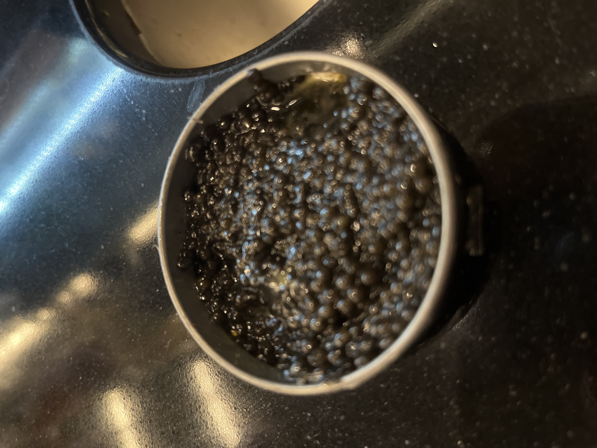 An overhead, close-up view captures a round, dark metal container filled with numerous small, glistening black spheres, likely caviar or fish roe, suspended in a dark liquid. The spheres have a uniform size and glossy appearance, reflecting ambient light. The container features a light-colored rim and sits on a dark, reflective surface, possibly a kitchen countertop or stovetop.
The surrounding surface exhibits a granular texture and strong reflections, showing diffuse patches of light and scattered bright highlights. In the upper-left portion of the frame, another circular depression or container, partially obscured, holds a lighter, opaque substance. The image is a still life, devoid of human presence or activity, and focuses entirely on the food item. The lighting suggests an indoor setting. No discernible text or specific branding is visible anywhere in the image. The scene is static and centered on the container of dark spheres.