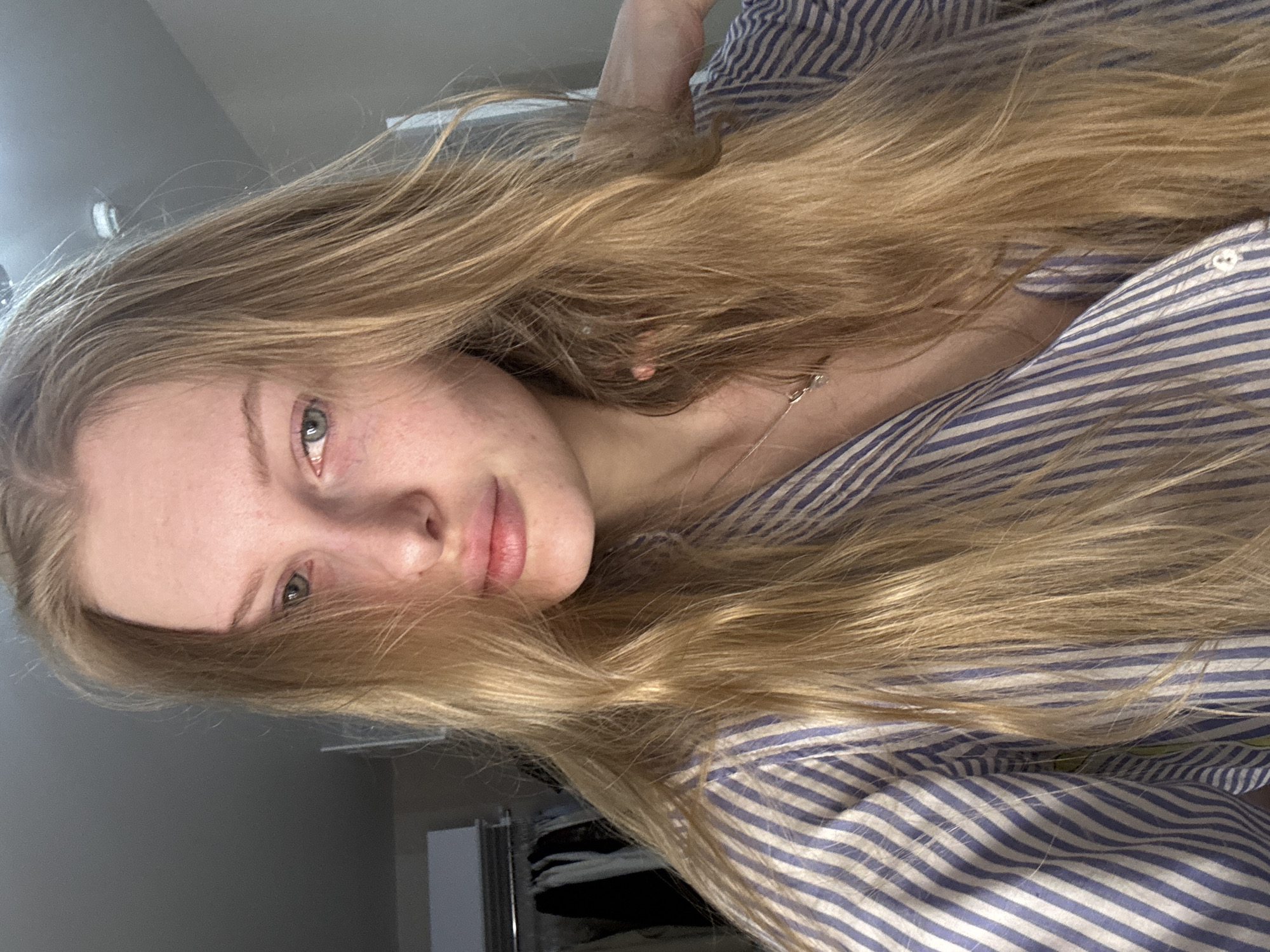 A young woman with long blonde hair and fair skin is lying down, possibly in a bed or on a soft surface, wearing a striped blue and white shirt. Her head is angled towards the viewer, with a slight tilt. Her eyes are green. A thin necklace is visible on her neck. The background appears to be a room with a wardrobe, possibly a bedroom. There's no evidence to confirm the specific location in Perm, Russia.