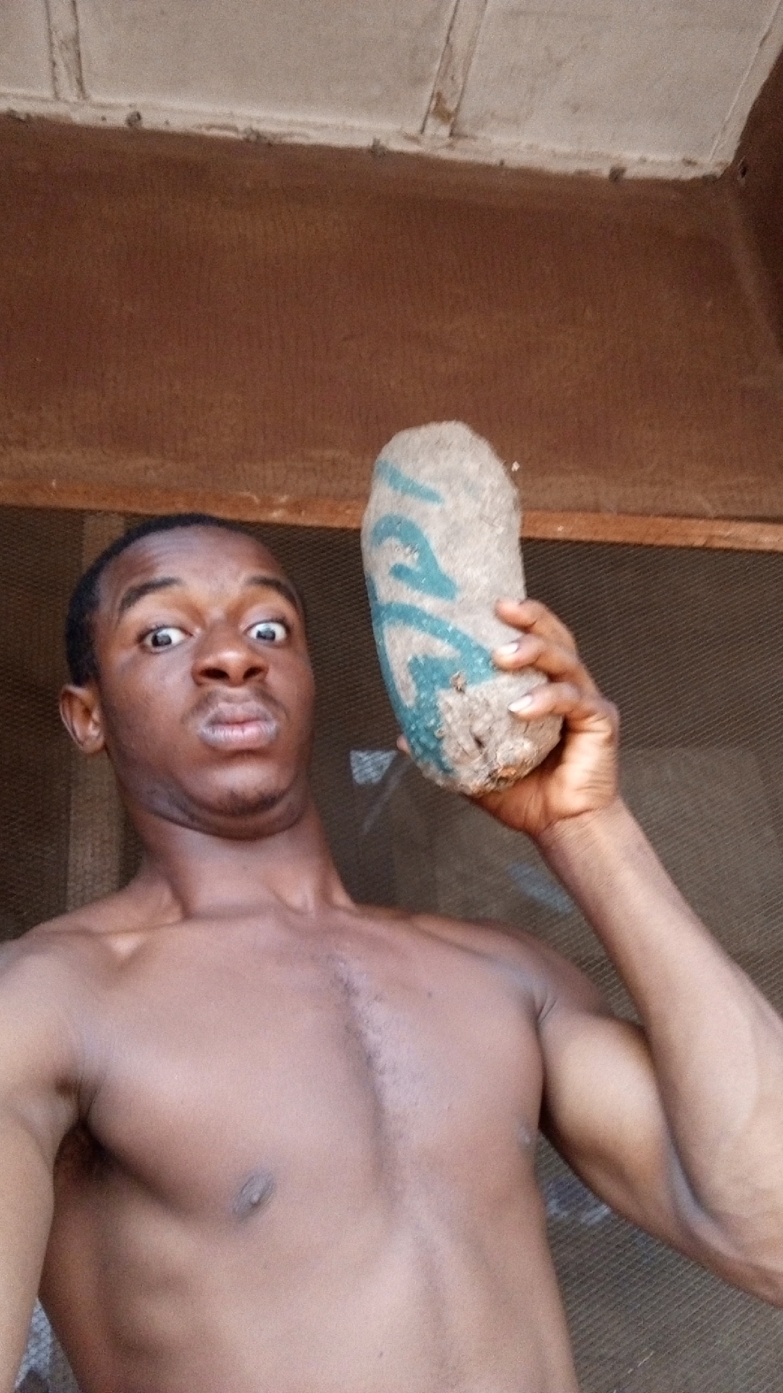 A shirtless, dark-skinned man with short black hair is holding a yam with green markings in his right hand. He is making a surprised expression. The background appears to be a wall or structure with a window. The location of the image is not clear from the picture. It is not possible to confirm the location is Onitsha, Nigeria from the image alone.