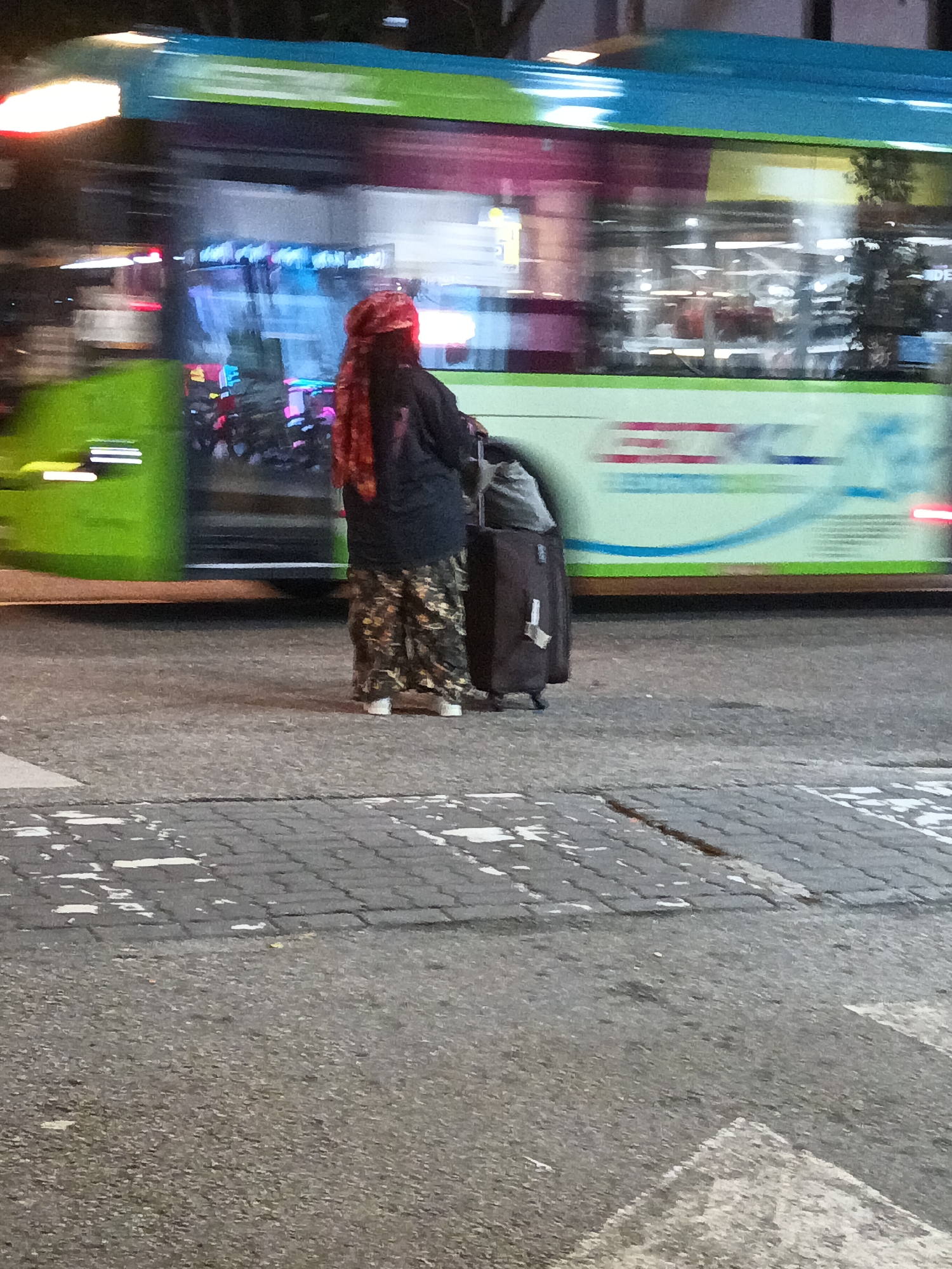 A person stands on a paved sidewalk next to a blurred green and blue bus. The person, facing away from the camera, wears a red head covering and patterned pants, and is holding a dark wheeled suitcase. The bus has windows and displays bright lights and colors, suggesting it is in motion. The ground is made of cobblestones and asphalt, with some white markings indicating a crosswalk. The scene is lit by artificial lights, suggesting it is nighttime or indoors. The image is blurry due to motion, making it difficult to discern specific details of the person's clothing or the bus's markings. Without more information, it is impossible to determine the specific location in Kuala Lumpur, Malaysia.
