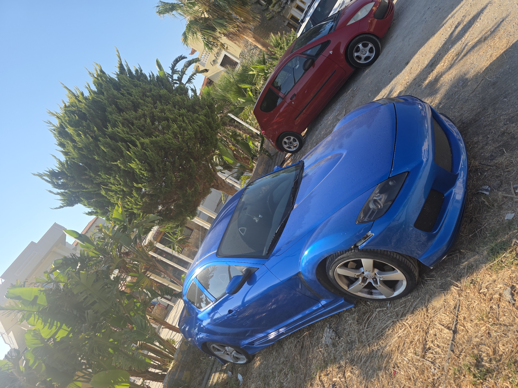 In the city of Malia Municipal Unit, Greece, on a sunny day, a blue Mazda RX-8 is parked on a dirt and dry grass surface. The car is angled towards the viewer's left, with its front facing slightly away. The blue paint has a metallic sheen, reflecting the bright sunlight. To the right and slightly behind the blue car, a red Mitsubishi Colt is parked, also at an angle. The Colt is a smaller hatchback. In the background, there are several large trees with lush green foliage, as well as a building with a light-colored facade and terracotta-tiled roofs. The sky is clear and blue. The scene depicts a street or parking area with vegetation and buildings typical of a Mediterranean climate.