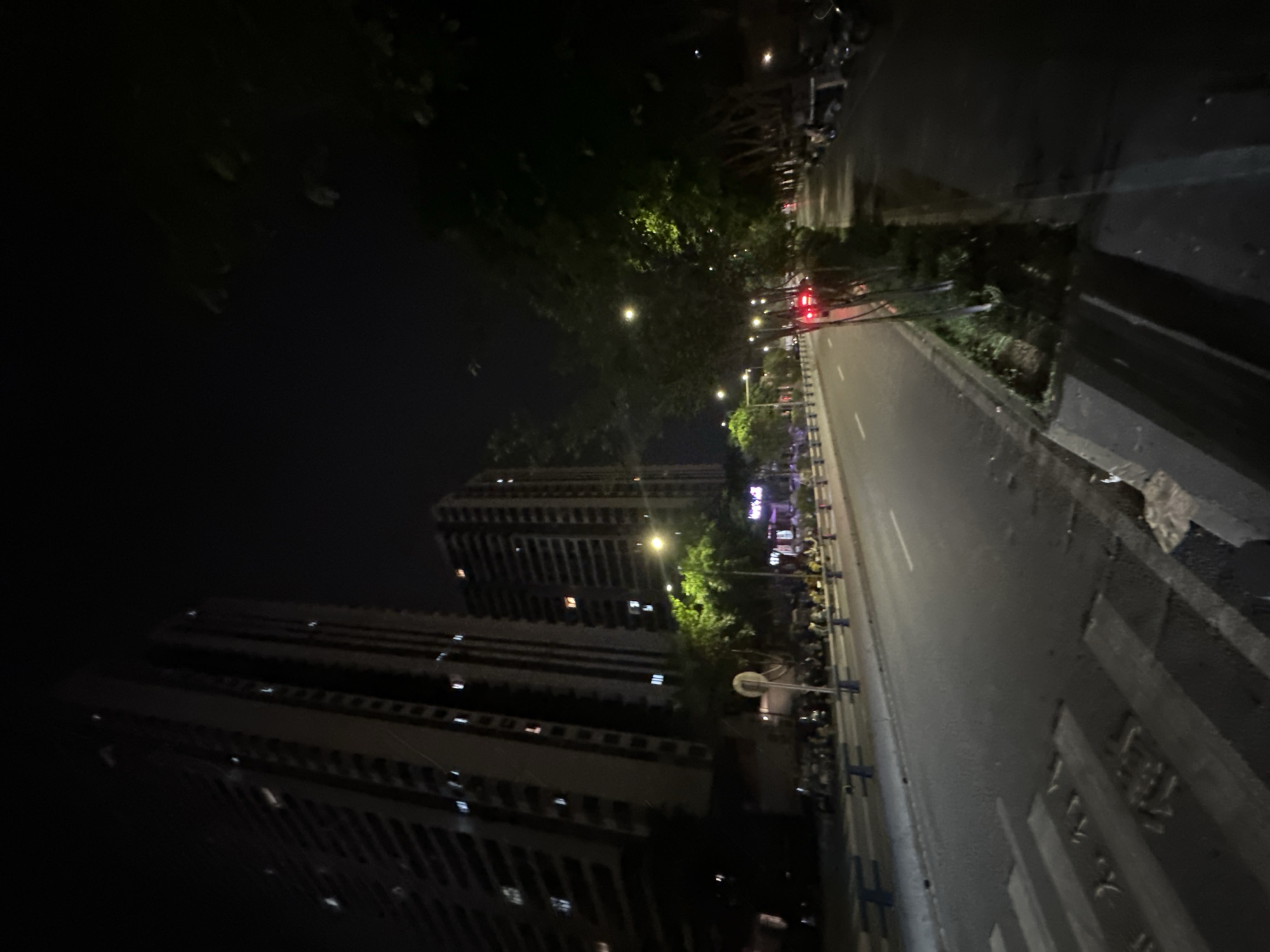 The image captures a nighttime street scene in the Chongqing High-tech Zone, China. Two tall, modern apartment buildings dominate the left side of the frame, their windows illuminated against the dark sky. A road with a pedestrian crossing is in the foreground, leading towards the buildings. On the right side of the road, trees with illuminated foliage line the sidewalk. Parked cars and motorcycles are visible on the far right, and a few people are gathered along the sidewalk. A red vehicle is seen driving on the road in the distance. The overall impression is one of an urban environment at night, with city lights and streetlights illuminating the scene.