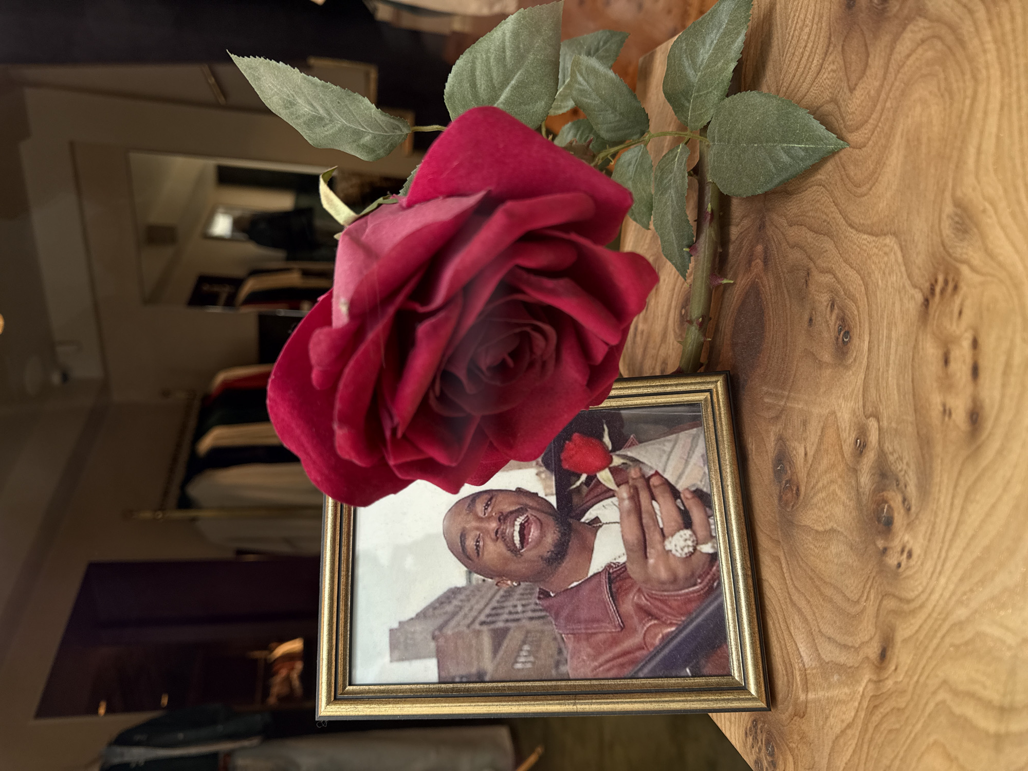 A framed photograph of Tupac Shakur holding a rose is displayed with a real red rose lying beside it. The frame sits on a wooden surface, likely a table or countertop. The photograph appears to be in a storefront window, with clothing racks and a mirrored wall visible in the background. No specific location in Paris, France, is discernible from the image.
