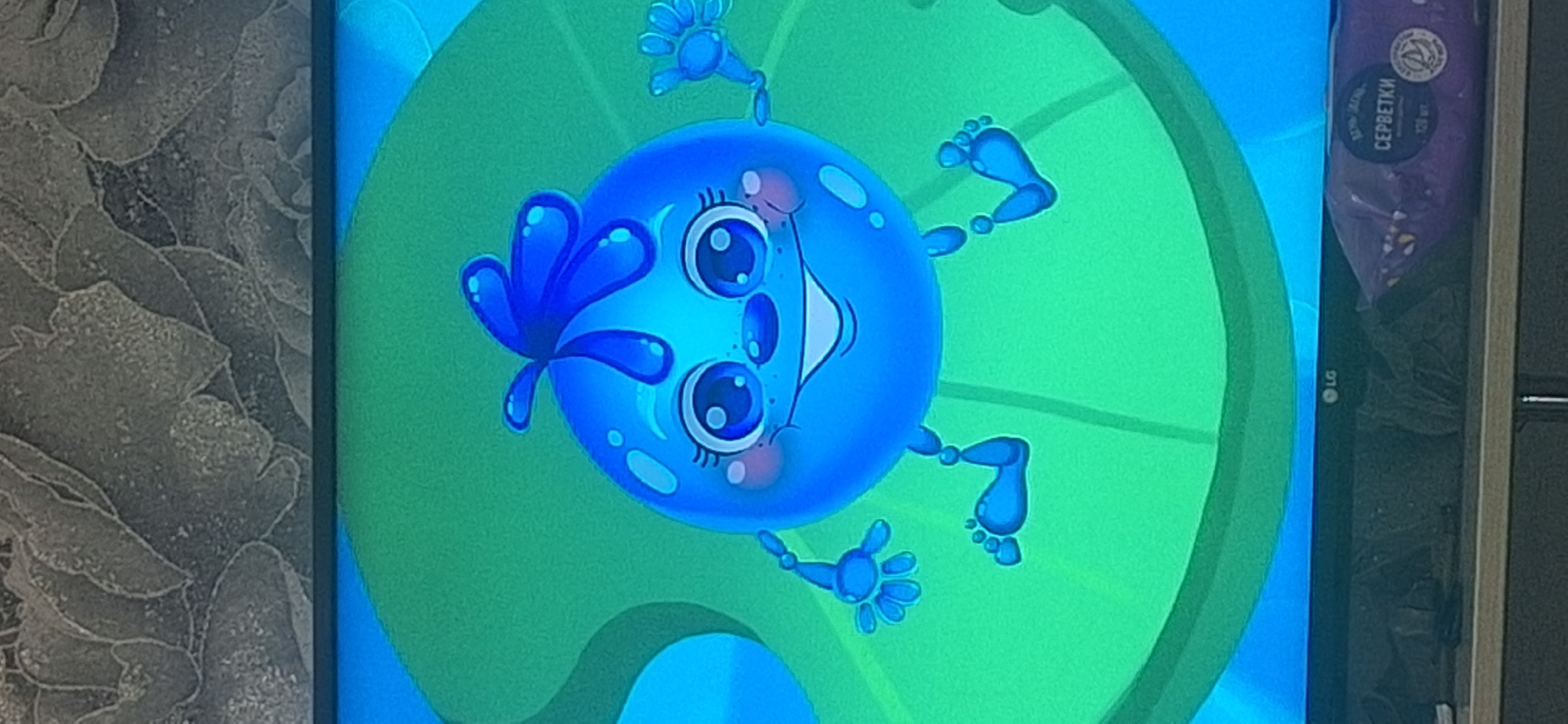 A digital illustration of a cartoon blueberry character is displayed on a television screen. The character is round and blue with large, expressive eyes, rosy cheeks, and a wide smile. It has thin, jointed limbs and what appears to be a leafy sprout on its head. The character is positioned against a bright green background with lighter green radiating circles. The bottom of the television screen shows a black bezel with an LG logo visible. Below the television, on a shelf, is a package of wet wipes with Russian text that translates to "WET WIPES" and a picture of a cat. The background above the television appears to be textured wallpaper with a grey, floral pattern. The overall scene is likely indoors, in a domestic setting, with no specific landmarks indicating Bila Tserkva, Ukraine.