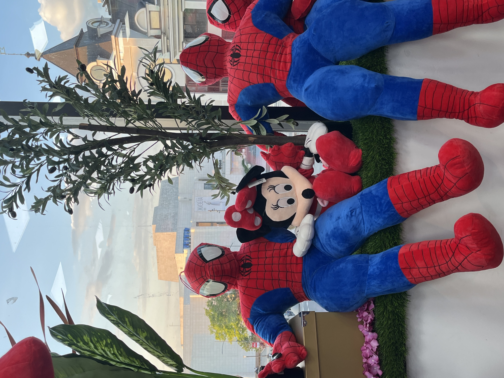 The image shows a display of plush toys arranged against a window. Two large Spider-Man plush toys, predominantly red and blue, are positioned adjacent to each other. A Minnie Mouse plush toy rests on one of the Spider-Man figures. The background is a cityscape reflecting in the window, with clear visibility of building facades and sky. Based on the architectural features visible in the reflection, the location is in Novi Sad, Serbia, and the building reflected is likely the Promenada Shopping Center. There are also artificial green plants framing the foreground. Artificial grass and purple flowers are also part of the display.