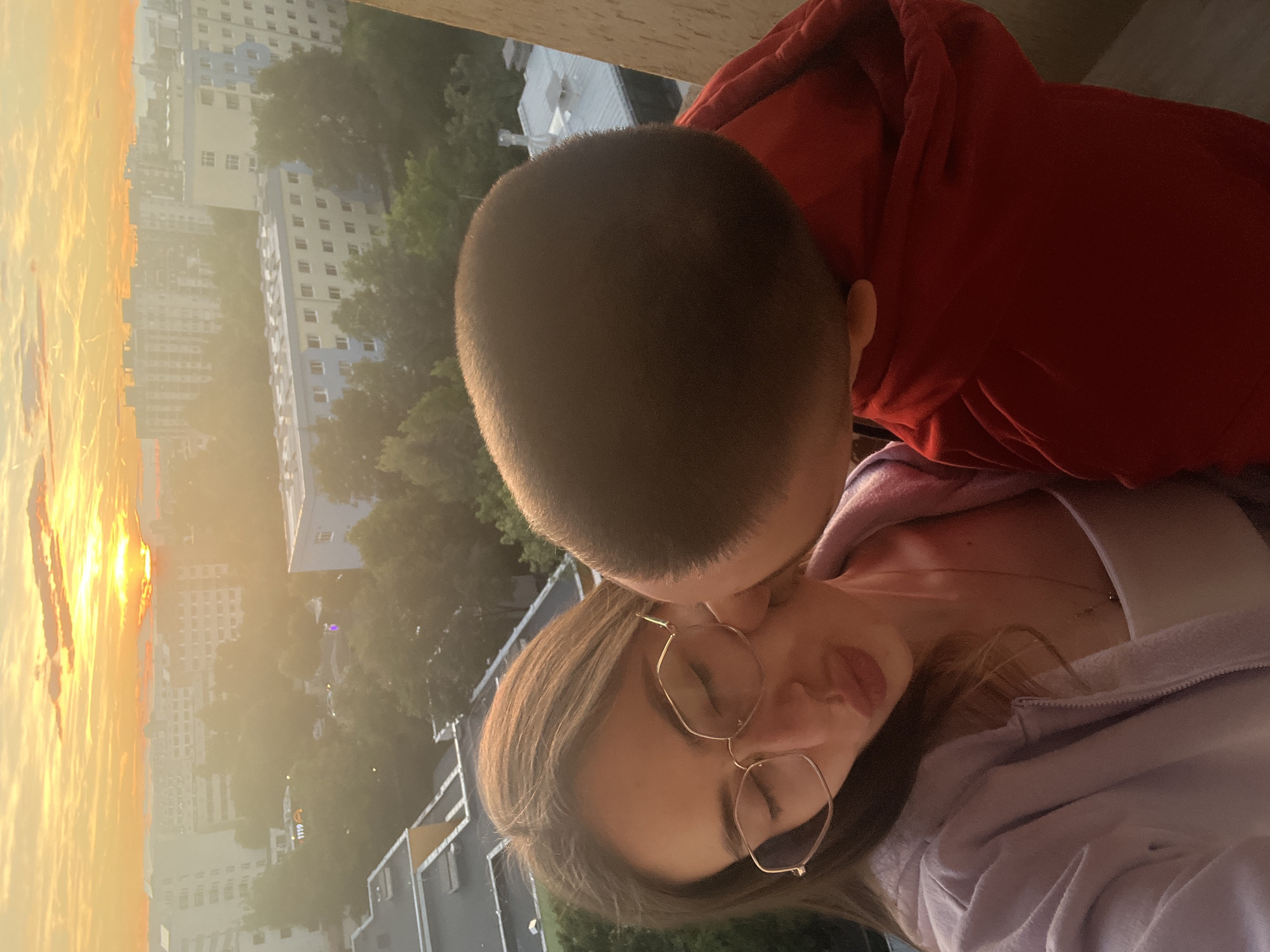 A medium close-up shot captures two individuals on a balcony at sunset, overlooking a city. The person in the foreground, on the left, is a young woman wearing glasses and a light purple zip-up hoodie over a white top. She has her eyes closed and her lips pursed as if to receive a kiss. To her right, a young child, wearing a red hoodie, is leaning in to kiss the woman's cheek. The background reveals a hazy cityscape with buildings and trees bathed in the warm, orange glow of the setting sun. The sky is filled with streaks of clouds and the faint outline of contrails.