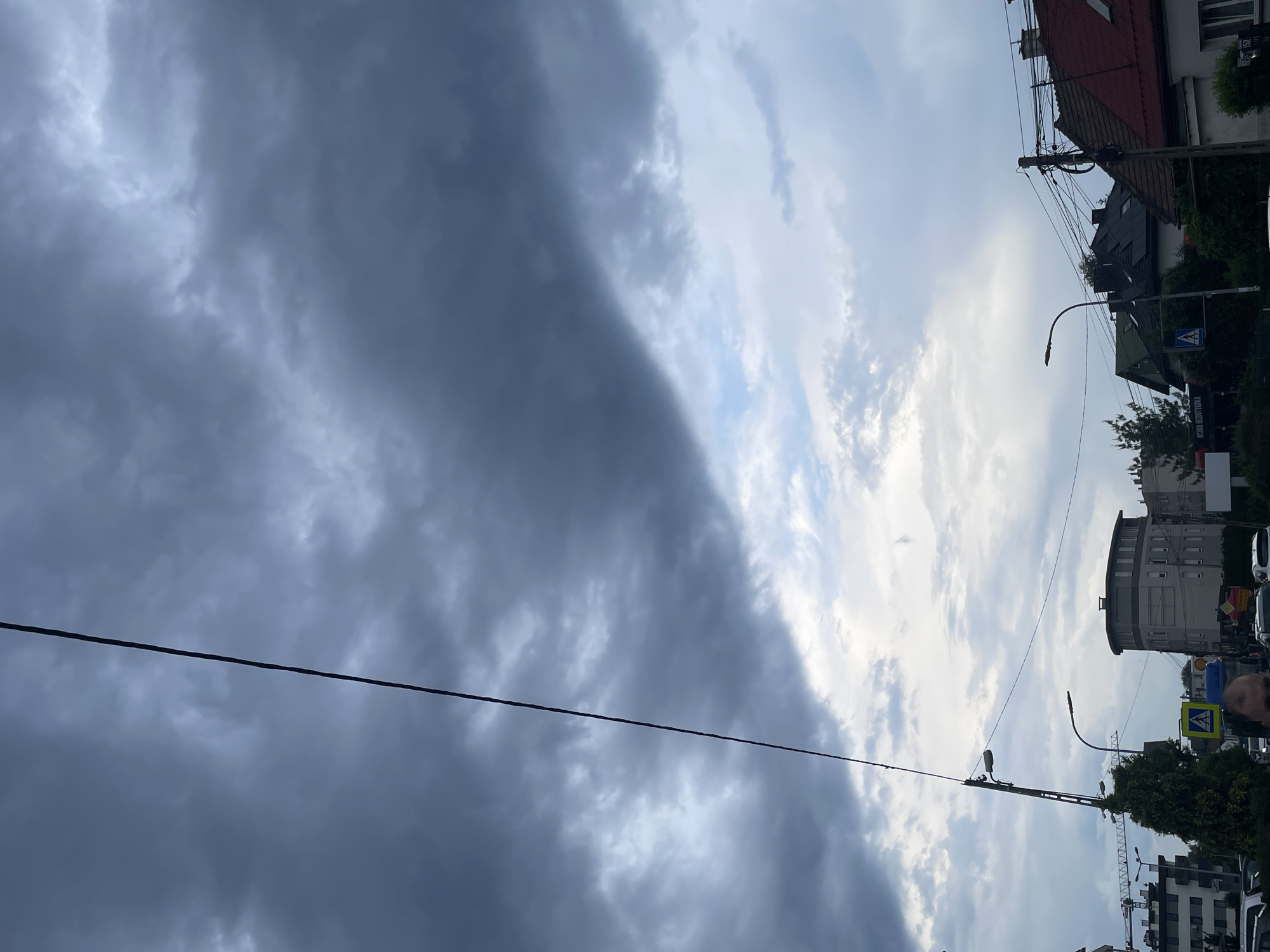 The image captures a dynamic urban or suburban scene, predominantly featuring a sky with stark weather contrasts. The left two-thirds of the sky are covered by heavy, dark gray clouds, indicative of an approaching storm or deeply overcast conditions. In contrast, the right third of the sky reveals lighter, brighter clouds with patches of blue sky, suggesting clearing weather or a localized weather front.
At the bottom of the frame, an urban environment is visible. On the right, several buildings line what appears to be a street. These include structures with red-tiled roofs and other multi-story buildings, one of which has a distinctive rounded, cylindrical section. Utility poles with numerous overhead wires crisscross the frame, with a thick black wire prominently stretching horizontally across the mid-section, visually dividing the darker and lighter sky areas. Two streetlights with curved arms are also visible.
Activity is suggested by the presence of partially visible vehicles (cars) and a construction crane in the bottom right corner, hinting at ongoing development. A yellow pedestrian crossing sign (blue triangle with a walking figure) is also seen on the right. A person's head and shoulder are partially visible on the far right edge of the frame, looking towards the left.
The time of day appears to be daytime, likely late morning or afternoon, given the overall illumination despite the heavy cloud cover. The most notable detail is the dramatic dichotomy of the sky, creating a sense of impending change in weather.
Visible text on a dark banner attached to a building on the right side includes the Polish word "WYNAJEM" (for rent/lease), suggesting commercial property is available.