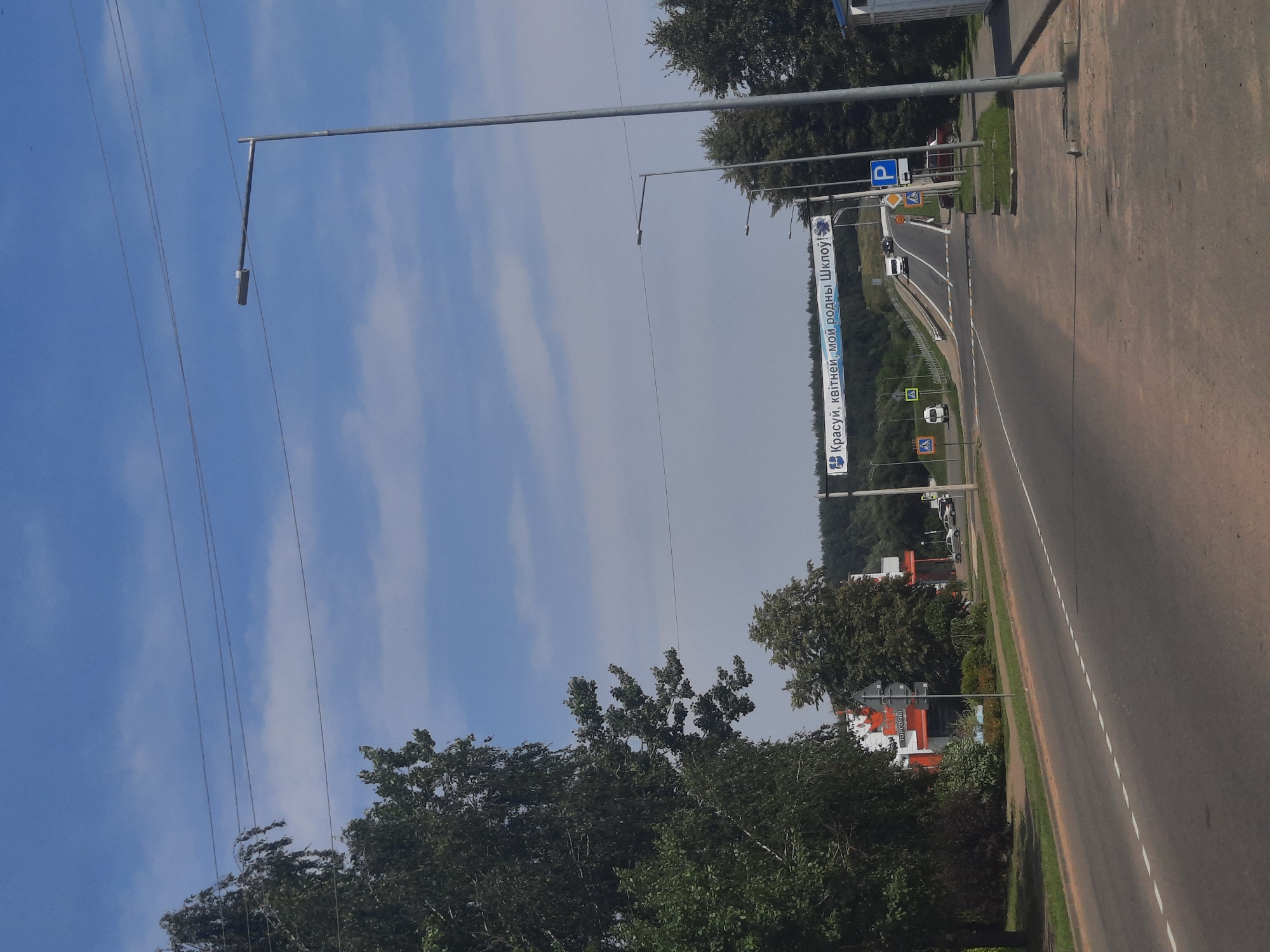 The image captures a sunny day with a bright blue sky and scattered white clouds, over a semi-urban or suburban area in Shklov, Belarus. In the foreground, a wide asphalt road with dashed white lane markings stretches into the distance, curving gently to the right. On the left side of the road, a dense cluster of green trees partially obscures a building with an orange and white facade, identifiable by Cyrillic signage that appears to read "ТОРГОВЫЙ" (TORGOVYI, meaning "Trade"). Power lines are strung across the upper portion of the frame.
Further down the road, a banner with Cyrillic text, "Красуй, квітней, мой родны Шклоў!" (Krasuy, kvitney, moy rodny Shklov!, meaning "Flourish, bloom, my native Shklov!"), is suspended across the road, indicating a welcoming message for the city. Several streetlights are visible along the roadside.
The road leads towards a slight incline, and several vehicles, including white vans and cars, are seen in motion. Road signs, including pedestrian crossing warnings and a blue parking symbol, are posted along the route. The background is dominated by a dense, dark green treeline of a forest. The overall scene depicts a typical day on the outskirts of a Belarusian city, with infrastructure and natural elements present.