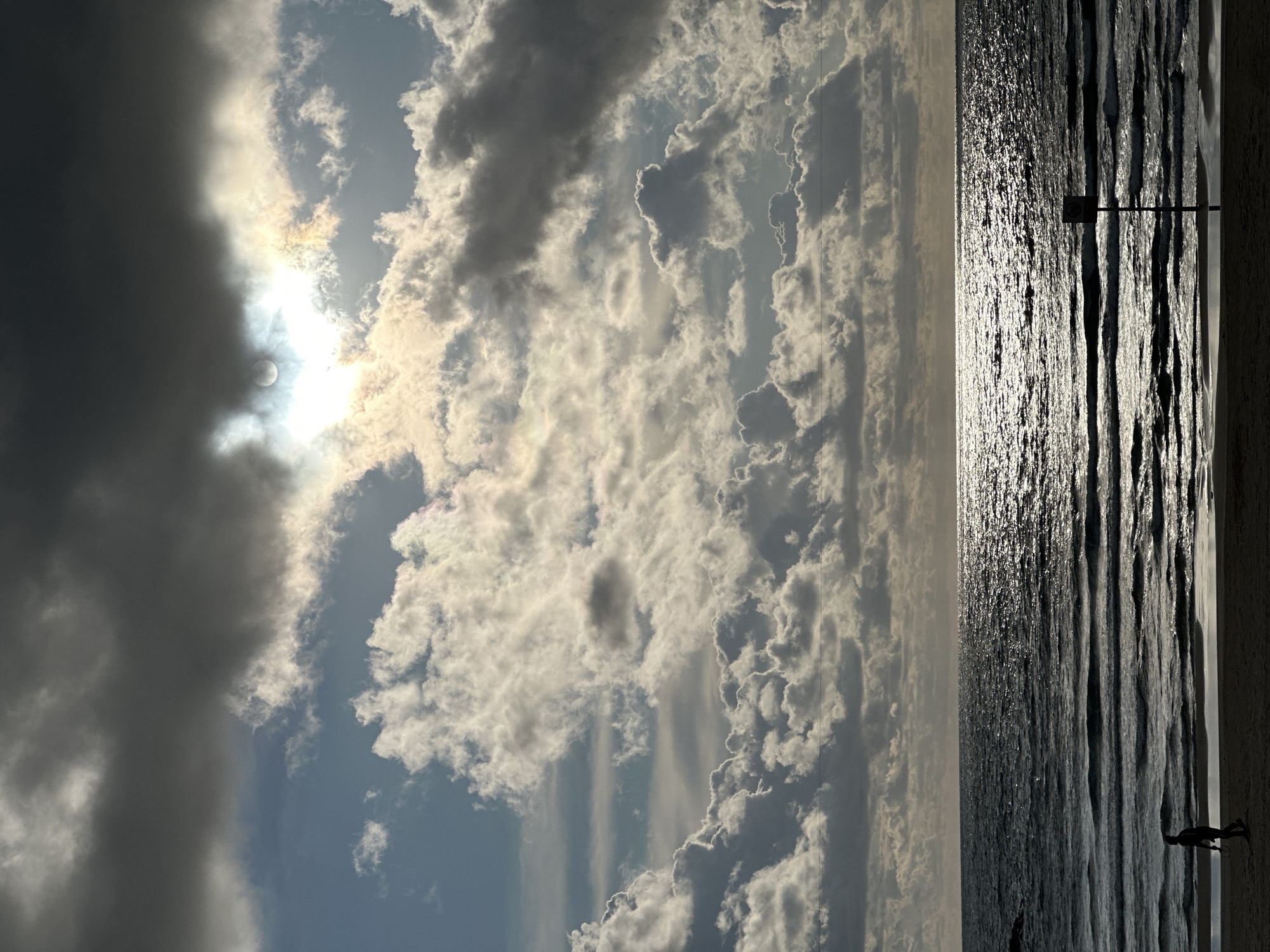 The image depicts a beachfront scene in Tel-Aviv, Israel, under a partially overcast sky. The sky, occupying the upper two-thirds of the frame, features a dense arrangement of cumulus and stratocumulus clouds. The sun, partially obscured by a large, dark cloud in the upper-center, emits bright light with a visible circular halo, casting iridescent reflections on surrounding clouds. Below the clouds, the Mediterranean Sea extends to the horizon, its surface marked by parallel waves. Intense specular reflection from the sun creates a shimmering, bright path across the water, particularly prominent in the mid-ground to the right. The water appears dark grey-blue. A narrow strip of dark, wet sand forms the immediate foreground. One solitary, silhouetted individual, appearing male and wearing shorts, walks from left to right on the wet sand near the water's edge in the lower-left quadrant. In the lower-right, a dark, slender pole with an indistinct, dark square or rectangular sign attached stands upright in the shallow water or wet sand. The overall scene is backlit by the sun, causing the human figure and the sign to appear in silhouette.