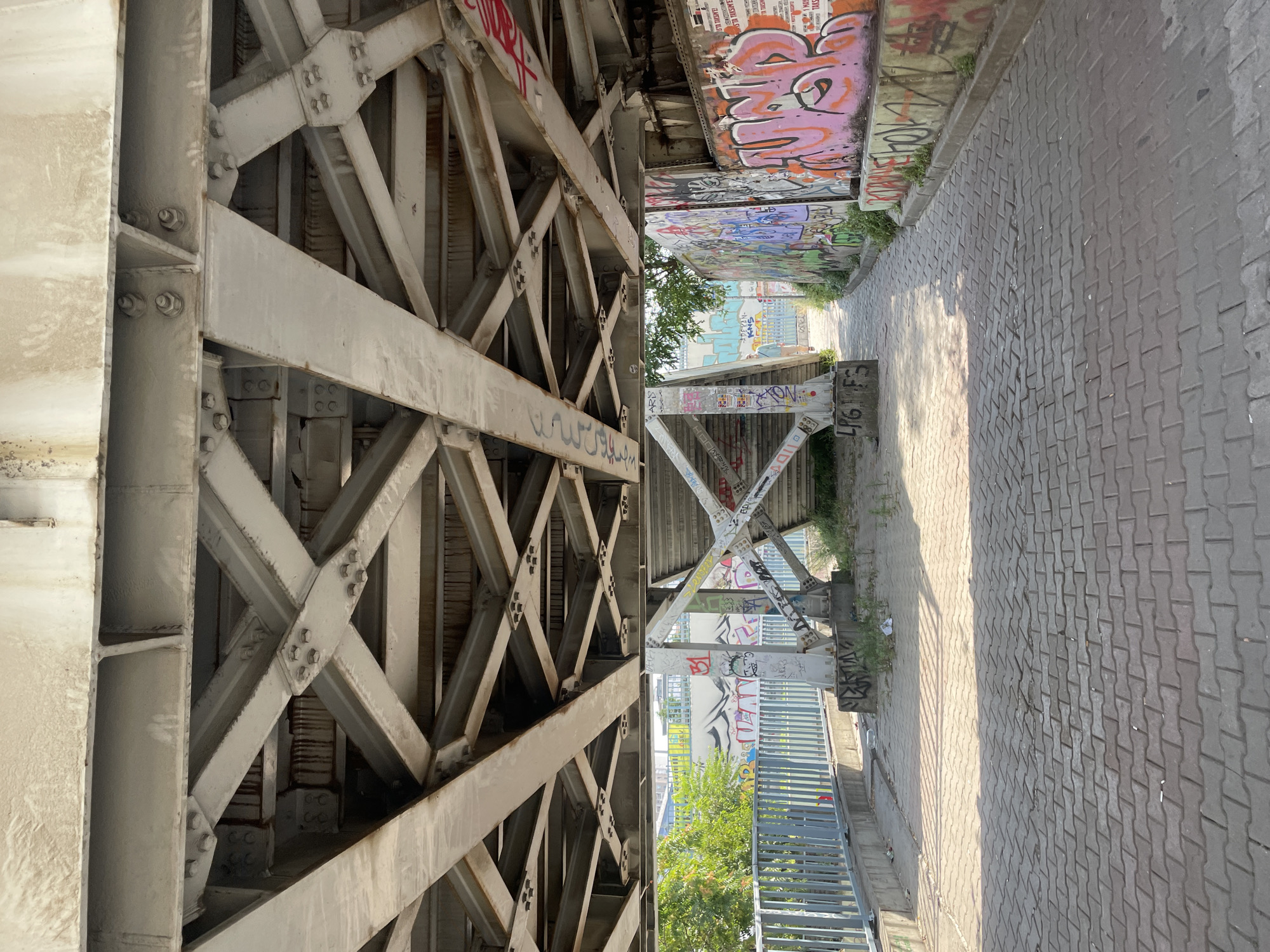 The image shows the underside of a large, grey metal bridge with intricate structural supports. The bridge's metal beams are crisscrossed and bolted together, showing signs of wear and some rust stains. Below the bridge, a paved walkway made of grey interlocking bricks runs through. To the left, a light blue metal fence borders the walkway, and beyond that, trees and more graffiti can be seen. To the right, a concrete wall is heavily covered in vibrant, colorful graffiti. In the distance, a person in a light blue top and dark pants is walking away from the camera on the walkway. Stairs leading up to the bridge are visible under the structure, also adorned with graffiti. The scene appears to be in an urban environment, likely a pedestrian underpass or a walkway beneath a bridge in Kadıköy, Turkey, given the distinctive graffiti style often found in the area and the general urban aesthetic. The sunlight casts distinct shadows on the brick pavement, indicating it is daytime.