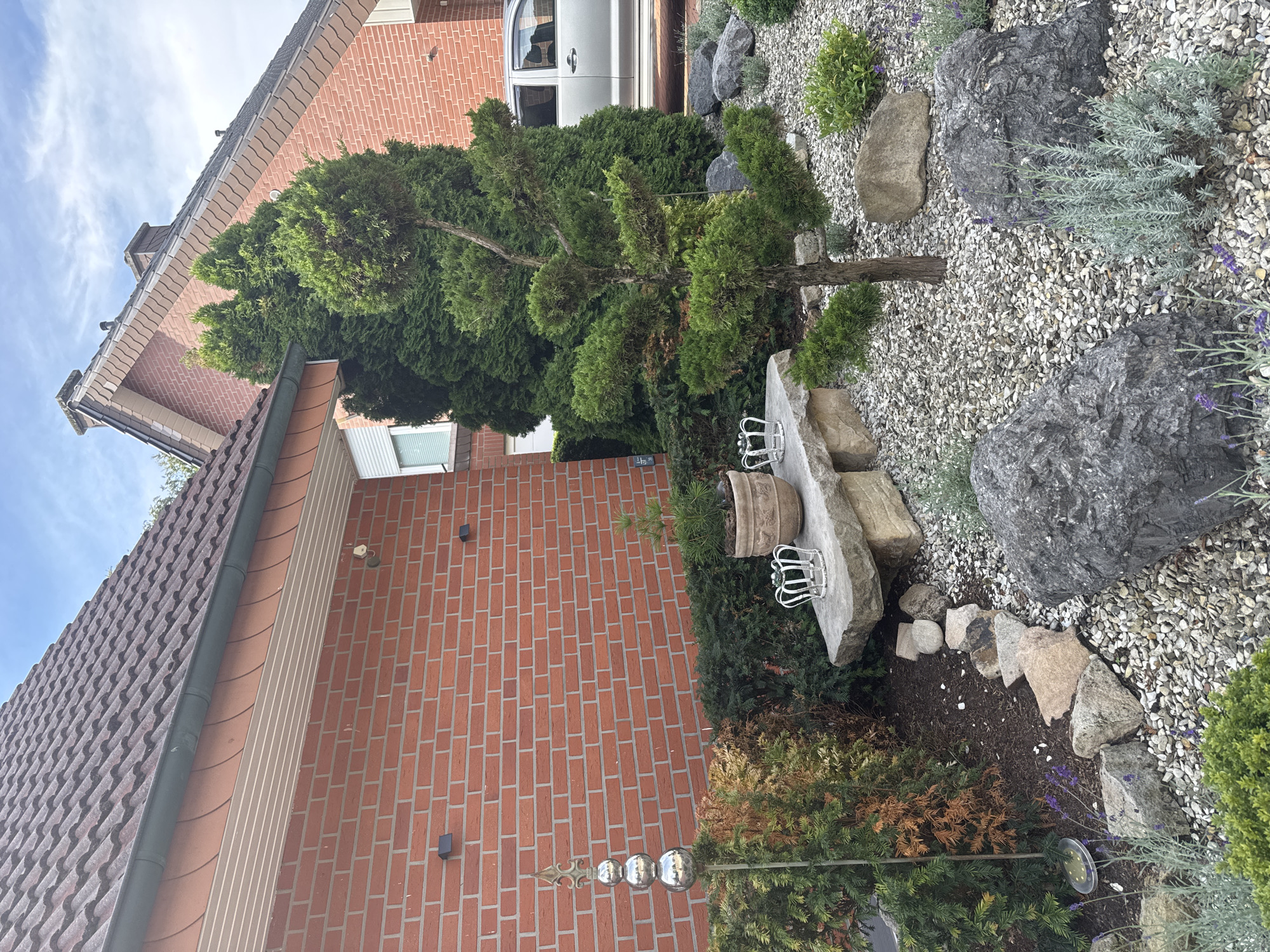The image depicts the front yard of a brick house in Rheine, Germany. The house has a red brick facade and a dark tiled roof. In the foreground, there is a meticulously maintained garden with a variety of plants, rocks, and decorative elements. A stone bench sits in the garden, adorned with two white crown-shaped ornaments and a pot with a small plant. A decorative metal stake with three spherical elements stands near the bench. The garden is landscaped with gravel, mulch, and several large rocks, along with low-lying shrubs and lavender plants. A portion of a silver van is visible parked in the driveway behind the garden. The sky above is partly cloudy.