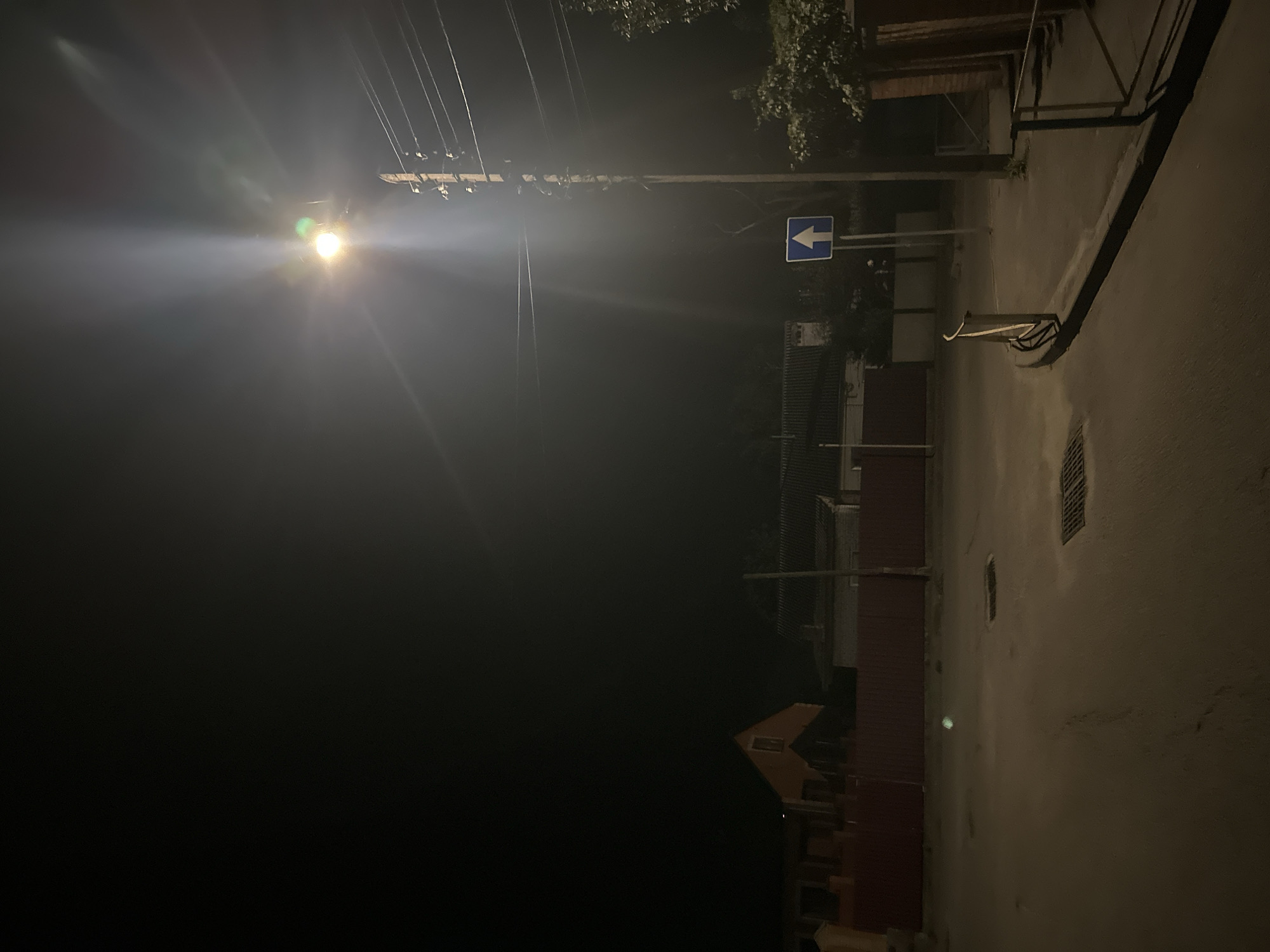 A nighttime street scene in Khmilnyk, Ukraine, is illuminated by a bright streetlamp. The lamp, positioned on a wooden utility pole on the right side of the image, casts a strong beam of light that creates lens flare and illuminates the surrounding area. The pole is adorned with insulators and a web of overhead wires. A blue square sign with a white upward-pointing arrow is mounted on a pole in front of the utility pole.
The street itself appears to be paved and slopes slightly downwards towards the left. Two rectangular metal grates are visible on the street surface. On the left side of the street, a red metal fence runs horizontally, bordering a property with a house that has an orange-tiled roof. Behind the fence, other structures with dark roofs are faintly visible in the dim light. To the right of the utility pole, a wooden structure with a sloped roof is partially visible, and a metal railing lines the edge of a pathway. Green foliage from a tree is present on the far right. The sky is dark, indicating it is nighttime.