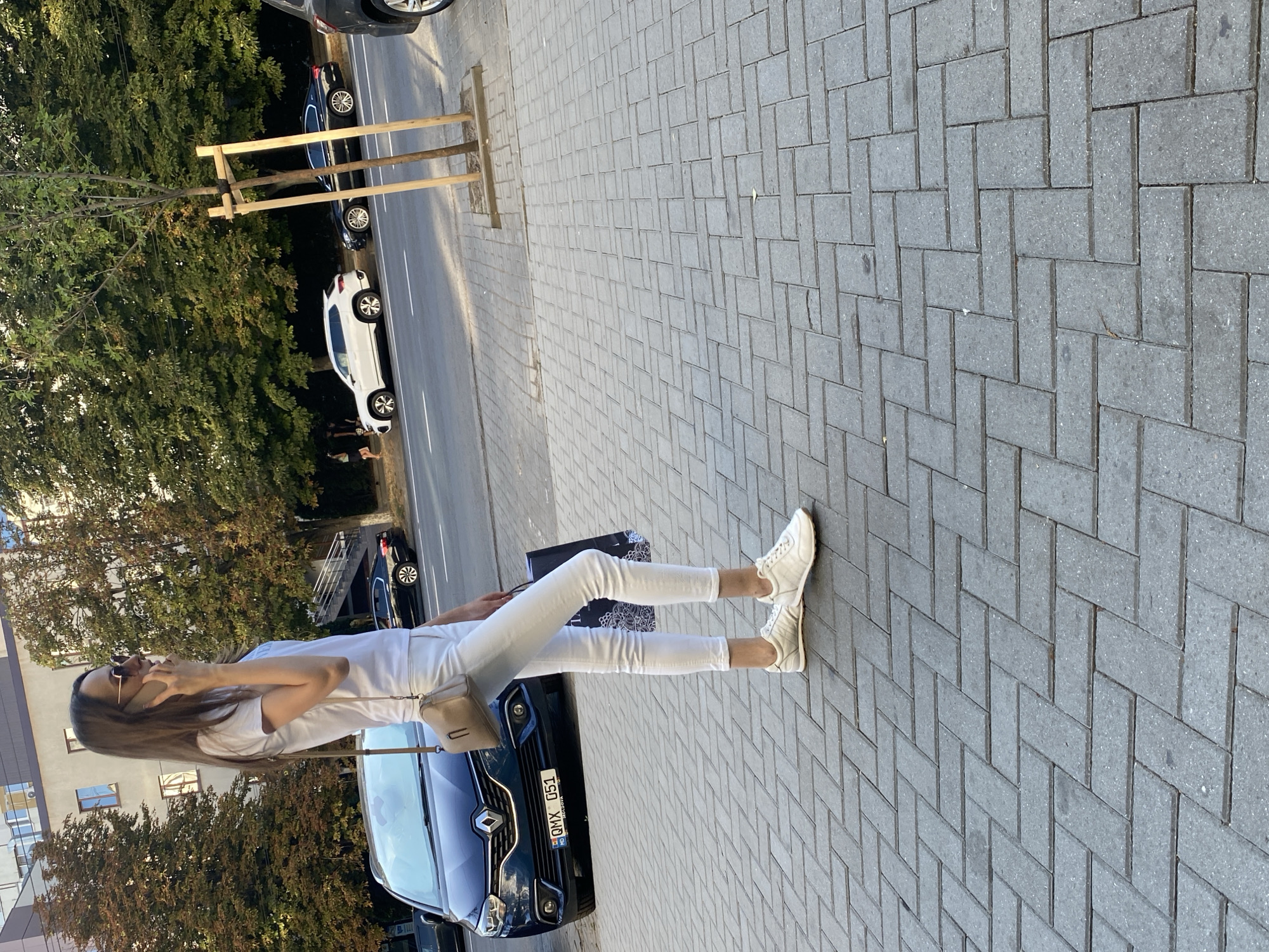 A woman stands on a cobblestone sidewalk in Chișinău, Moldova, talking on her phone. She is wearing white skinny jeans, a white t-shirt, and white sneakers. A beige crossbody bag hangs from her shoulder, and she carries a dark shopping bag in her right hand. The woman is looking to her right. Behind her, a dark blue car with a license plate reading "QMX 051" is parked. In the background, trees line the street, and several cars are parked along the curb. The scene appears to be a street in a city during the daytime, with sunlight casting shadows on the ground.
