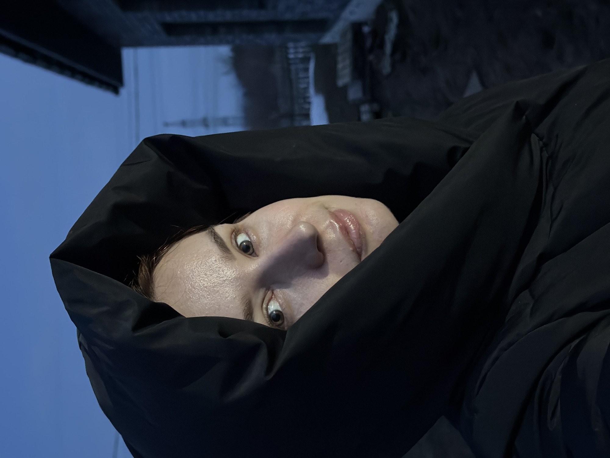 A person is seen in a close-up, angled portrait, predominantly obscured within the hood of a dark jacket. The individual has fair skin, slightly visible brown hair, and a light gloss on their lips. Their eyes are wide open, gazing directly at the camera.
The backdrop indicates an outdoor setting in the evening or early morning. Above the person, there are blurred hints of a building structure, possibly part of a cityscape, and overcast sky. The exact location in the city of Chaikovsky, Russia, cannot be definitively determined from this image alone.