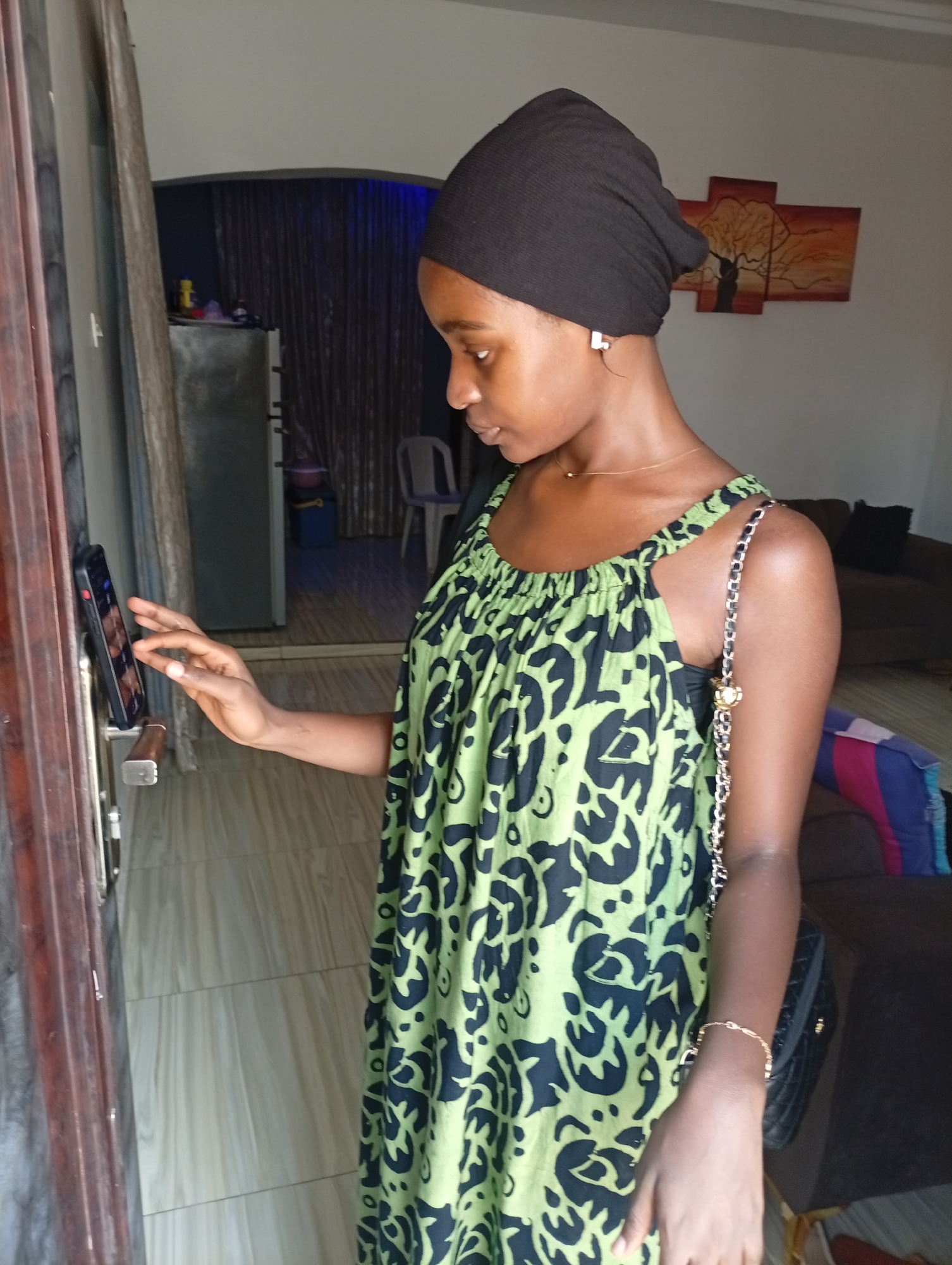 A young woman wearing a black head covering and a green and black patterned dress is standing in a doorway, possibly to her home. She is touching a smartphone attached to the door. A silver chain purse hangs over her shoulder. Inside the doorway is a tiled floor, a refrigerator, and a painting of a tree on the wall. The location is Unknown, Unknown.