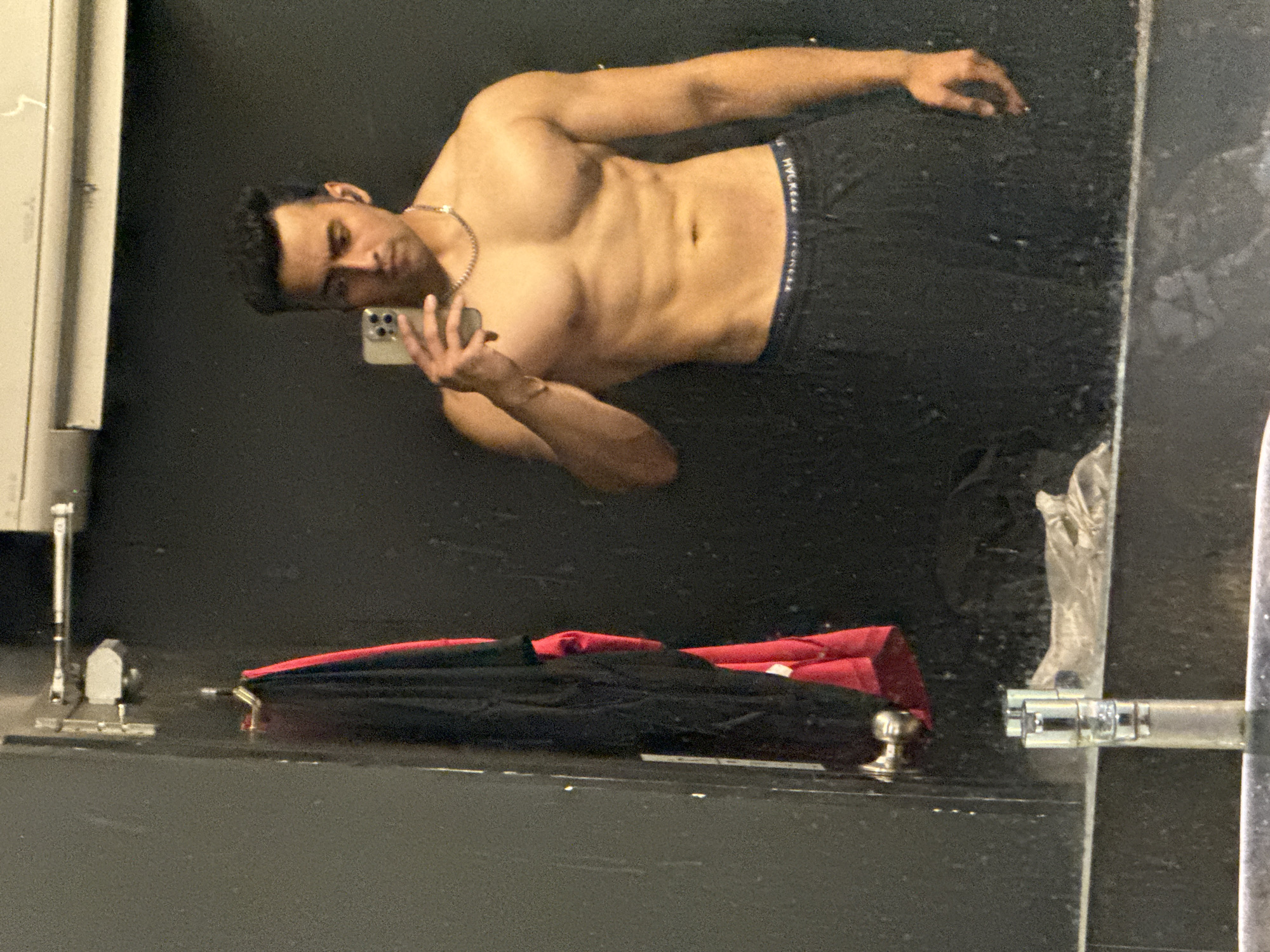 A man is taking a selfie in a mirrored wall in a room. He is shirtless and wearing dark-colored bottoms. His right arm is outstretched, holding a mobile phone. He has dark hair and is wearing a chain around his neck. On the shelf below the mirror, there is a black and red folded umbrella, as well as some other indistinct items. The location is undetermined as the image contains no elements that are specific to Kolkata, India.