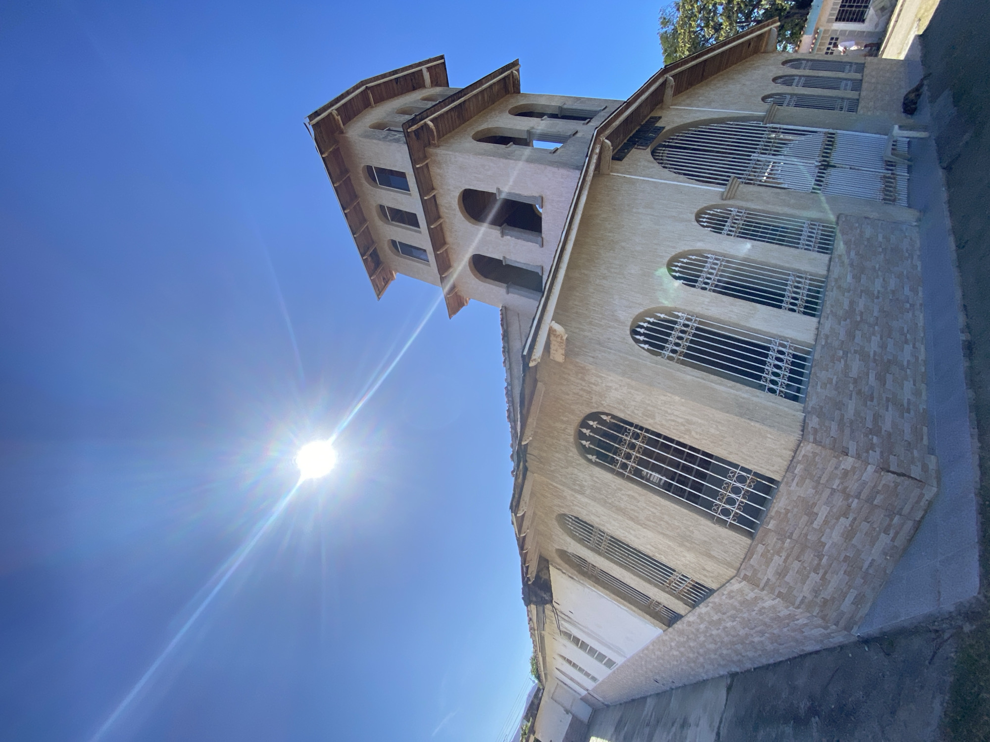 The image shows a tall, light-colored building with a tower under a bright blue sky in Juan Griego, Venezuela. The sun is shining intensely, creating glare. The building has arched windows with white metal grilles. The tower has multiple levels with arched openings, some of which are open to the outside. The lower portion of the building has tile accents. The architecture is simple and somewhat weathered. A sidewalk runs along the base of the building. The perspective is a low-angle shot looking upwards towards the building and sky.