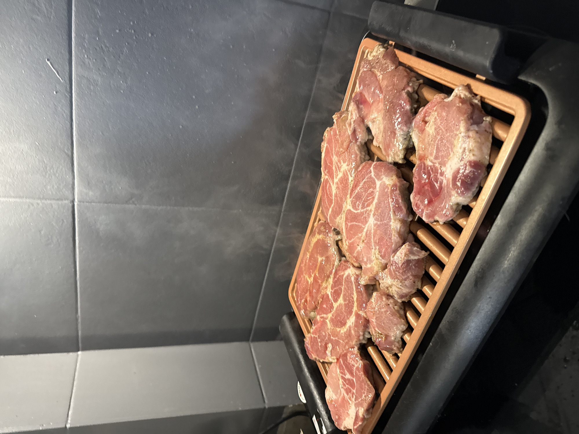 The image displays an indoor cooking scene, focusing on an electric grill laden with numerous pieces of raw, seasoned meat. The meat, possibly pork neck or shoulder, exhibits a reddish-pink hue with distinct marbling and appears to be coated with spices, indicating preparation for grilling. The grill features a copper-toned non-stick grate set within a black, industrial-looking base. A faint haze or steam is visible above the grill, suggesting it is either preheating or has just commenced cooking.
The background consists of dark grey, possibly ceramic, tiled walls, characteristic of a modern kitchen or a dedicated cooking space. The lighting in the scene is artificial, illuminating the grill and meat clearly. No individuals are present, and there is no visible text. The image captures the initial stage of a meal preparation, highlighting the raw ingredients and cooking equipment. The time of day and external weather conditions are not discernible from this interior shot. The overall impression is one of anticipation for a grilled meal.