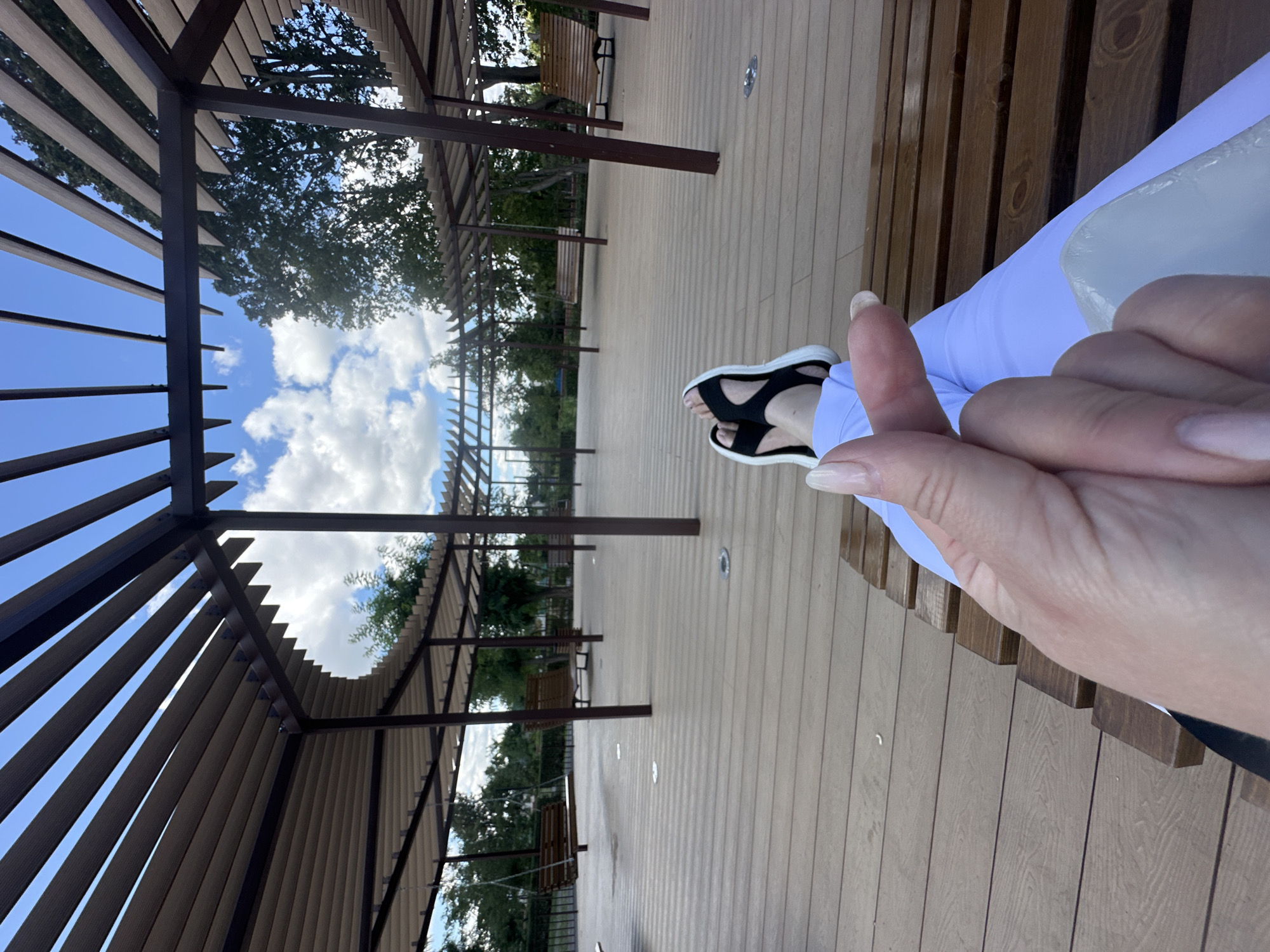 The image depicts an outdoor seating area with a wooden deck, covered by a slatted canopy structure. In the foreground, a person's legs and hands are visible, with the person wearing white pants and black and white sandals. They are sitting on a wooden bench. In the background, there are more benches and a swing, set against a backdrop of trees and a blue sky with white clouds. The scene appears to be a public park or recreational space, potentially located in Zhovte, Ukraine, though this cannot be definitively confirmed from the image alone.