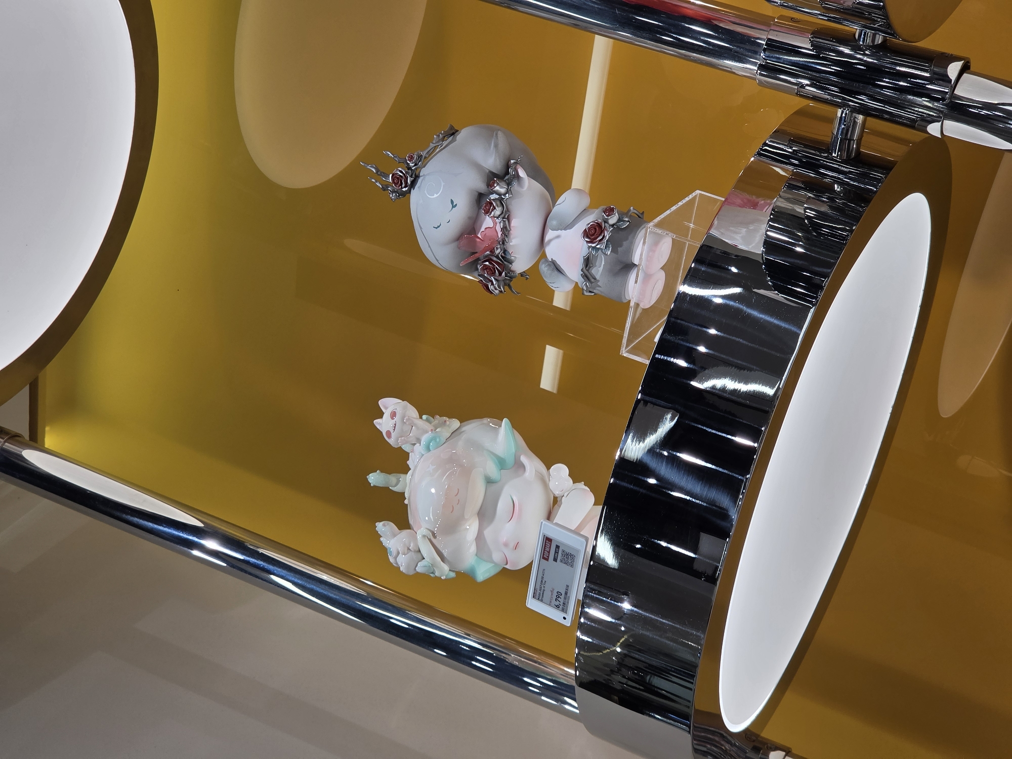 Two collectible figurines are displayed in a showcase, likely for sale. The lower figurine is white and pink, decorated with small cats. The higher figurine is grey and pink, decorated with roses. A price tag is visible near the lower figurine, indicating a cost of 6,790. The showcase is constructed from chrome metal and yellow-gold colored acrylic. The photograph was taken in Bangkok, Thailand.